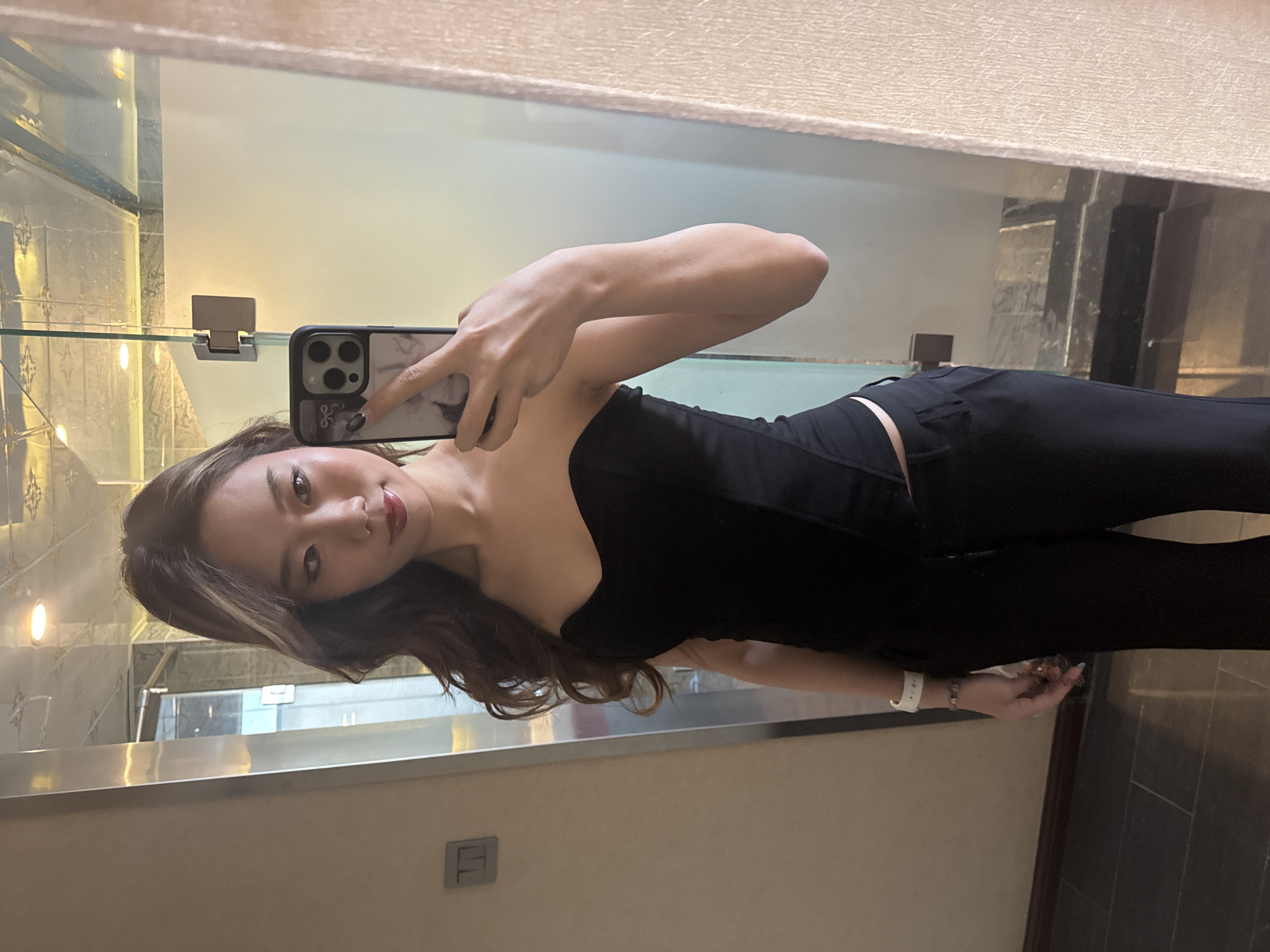 A young East Asian woman is captured in a mirror selfie, posing in an indoor setting in Ganzhou, China. She has long, wavy dark brown hair with lighter streaks, styled over her shoulders. Her face is made up, featuring dark eyes and red or pink lipstick, and she has a calm, slightly smiling expression as she looks into the camera.
She is dressed in a black, off-the-shoulder or strapless top, possibly a corset or bustier style, paired with form-fitting black pants. On her right wrist, she wears a white smartwatch and a beaded bracelet. She holds a dark-colored smartphone, likely an iPhone, in her left hand, which is encased in a patterned, light-colored cover featuring a small black bow or ribbon design near the bottom.
The background, visible as a reflection in the mirror, suggests a modern and well-lit interior. There's a light-colored, textured border at the top and bottom of the mirror. On the left side of the reflection, a glass shelf with a metallic bracket is visible, with light-colored, possibly patterned tiles behind it. Further into the reflection, there are darker, potentially gray or black wall panels. A circular light fixture can be seen reflected on the left. The overall ambiance suggests a contemporary space, perhaps a bathroom, dressing room, or hallway in a building. The woman appears confident and relaxed while taking the photograph. No discernible text is visible in the image.