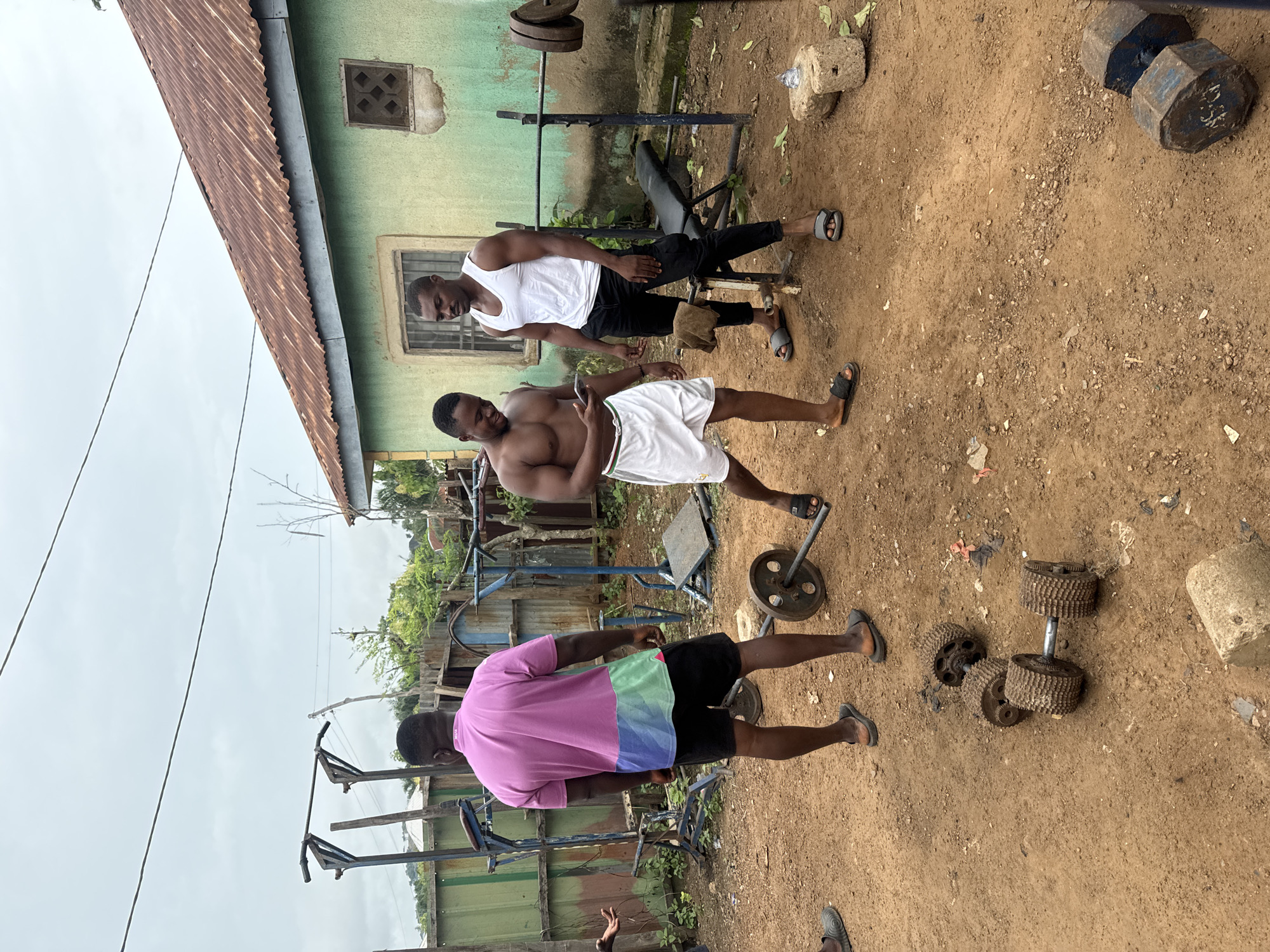 Three men are gathered in an informal outdoor gym setting on reddish-brown dirt ground, likely in Akwanga, Nigeria. The sky is overcast, suggesting daytime.
The subjects include three adult Black males. One man stands shirtless, wearing white shorts and black sandals, holding a smartphone in his right hand. Another man, wearing a white tank top, black trousers, and grey sandals, stands next to a metal bench press with a barbell loaded with circular weights. The third man, with his back mostly to the camera, wears a multi-colored t-shirt (pink, light green, light blue sections), black shorts, and black sandals.
The scene features various pieces of exercise equipment scattered across the dirt. These include the metal bench press, several barbells, and dumbbells. One octagonal dumbbell weight clearly displays "25KG". Some dumbbells appear to be crafted from repurposed gears. In the background, a single-story building with a light green painted wall and a rusted corrugated metal roof is visible. A wooden vent is set into the wall, and parts of a window with metal bars can be seen. Sparse vegetation and overhead power lines are also present.
The men appear to be either working out, taking a break, or socializing at this rustic gym. Their demeanor seems relaxed and casual.