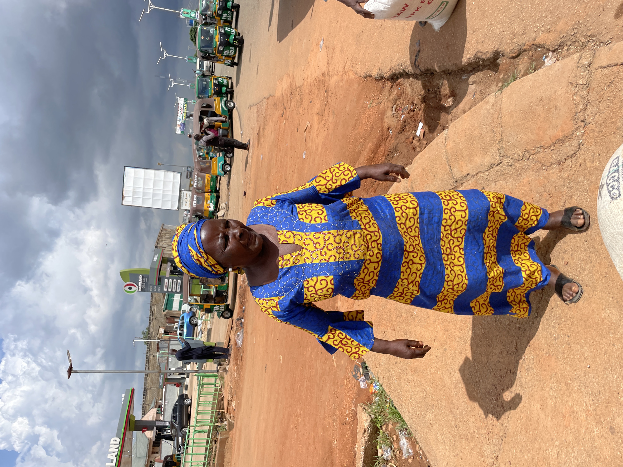 In Gura Top, Nigeria, a woman stands in the foreground, her blue and yellow patterned dress and headscarf vibrantly contrasting with the dusty, reddish-brown ground. She wears black sandals. Behind her, a line of green and yellow tuk-tuks, common modes of transport in the region, are parked along a dirt road. A white billboard stands prominently in the mid-ground. Further back, a gas station with a prominent "MARILAND Oil & Gas" sign and pricing details for PMS, AGO, and DPK is visible. The sky is cloudy, suggesting a potentially overcast day, but the overall lighting indicates it is daytime. The scene captures a typical street view in a Nigerian town, with people, vehicles, and businesses coexisting. The woman's gaze is directed slightly upwards, her expression neutral. The image conveys a sense of daily life and commerce in this Nigerian location.