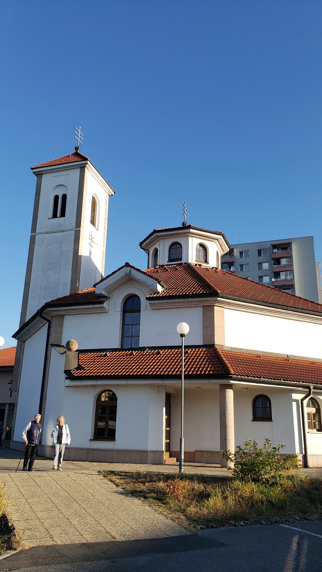 In the city of Prešov, Slovakia, a white church with a red-tiled roof stands under a clear blue sky. The church features a tall, narrow bell tower with a cross at its peak. A smaller domed section with arched windows is situated on the main building, also topped with a cross. To the right of the church, a multi-story apartment building is visible. In the foreground, two elderly individuals, a man with a cane and a woman, walk on a paved walkway. The ground is a mix of pavement and dry grass. The lighting suggests it is daytime, likely late afternoon due to the long shadows.