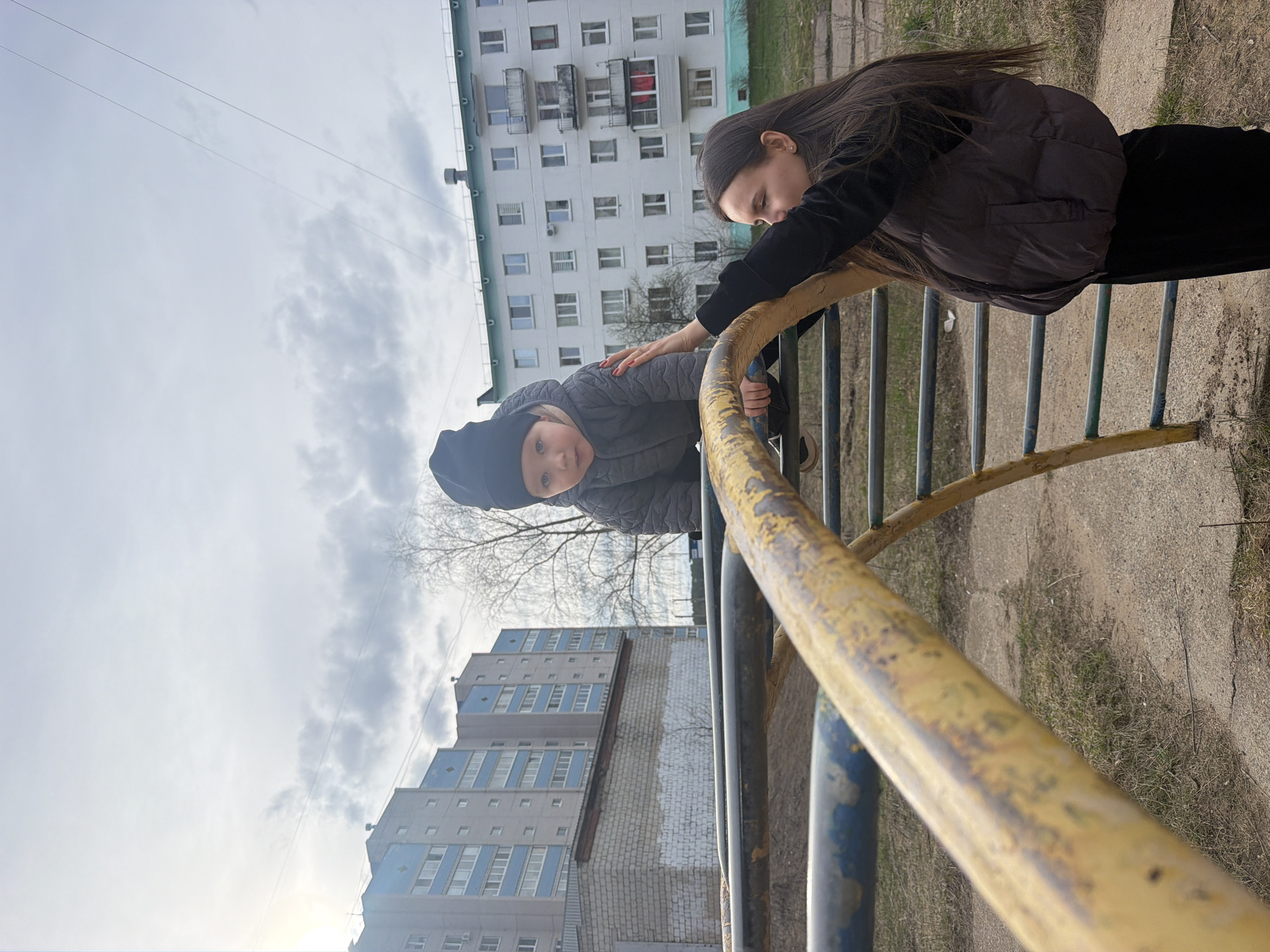 An image captured in Chaikovsky, Russia, shows an adult and a child on a playground, viewed from a low, upward-looking perspective. The photograph is oriented vertically, suggesting it was taken with the camera rotated.
In the foreground, a yellow and partially blue metal playground structure, possibly monkey bars or a climbing frame, dominates the lower half of the frame. An adult, a woman with long, dark hair, is seen leaning over the yellow bar, her right hand gently placed on the back of a small child. She is wearing a dark long-sleeved top and a dark, sleeveless puffer jacket.
The child, who appears to be a toddler or preschooler, is wearing a dark beanie and a grey puffer jacket. The child's face is turned towards the camera, peeking over the yellow bar with a direct and curious gaze.
In the background, behind the playground structure, several multi-story apartment buildings are visible under a cloudy sky. One prominent building is white with numerous windows and balconies, characteristic of Soviet-era architecture. Another building, further back, features blue and grey sections with multiple windows. Bare trees, indicative of late winter or early spring, are also visible between the buildings and the playground. The ground beneath the playground is a mix of dirt and sparse, dry grass.
The scene depicts an interactive moment between the adult and child on a cloudy day, likely on a public playground in an urban residential area. No visible text is present in the image.