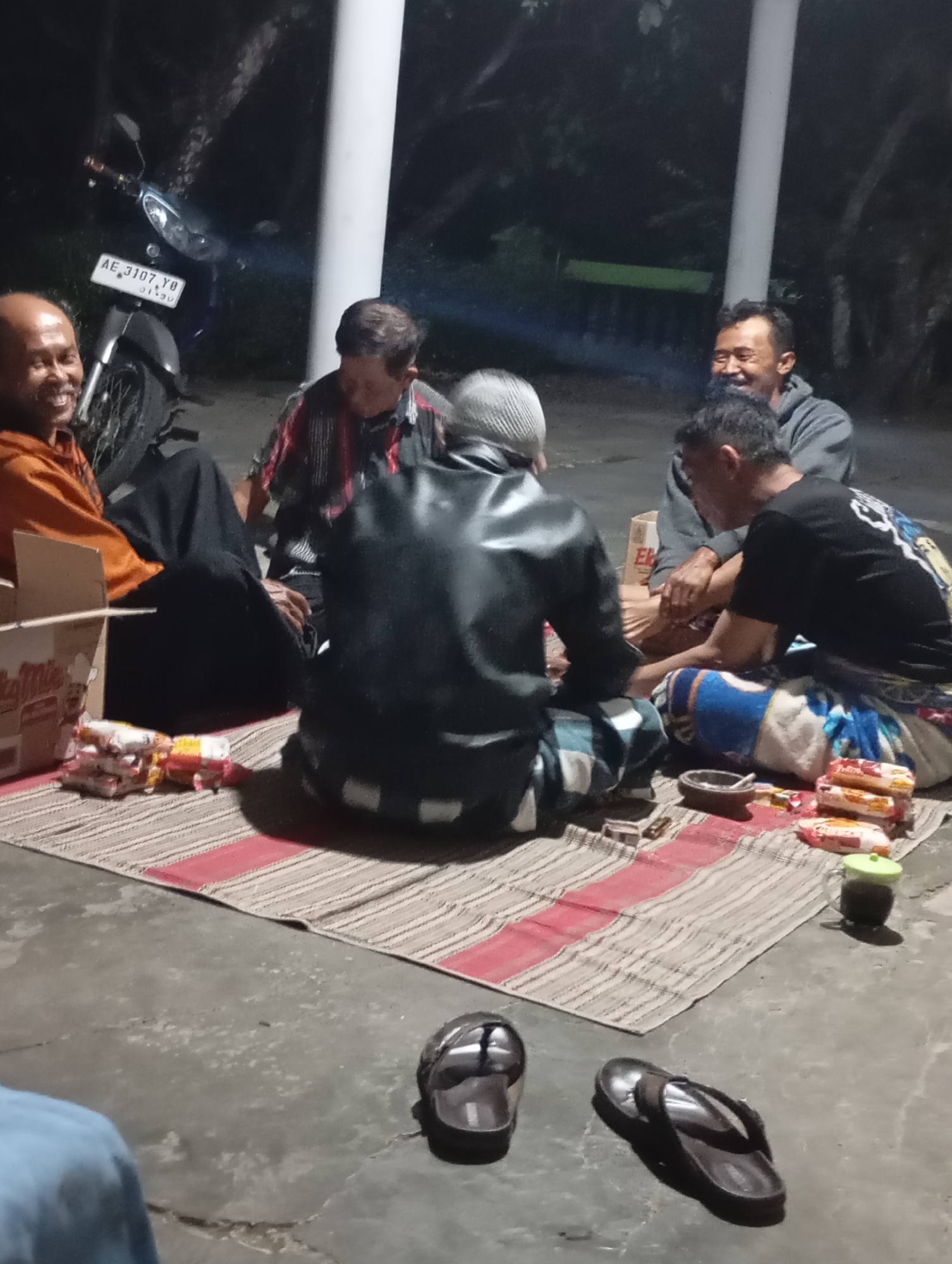 Six adult males are gathered outdoors on a paved surface, sitting on a woven mat. They appear to be in a casual, social setting. One man on the far left, wearing an orange shirt, is smiling broadly. Next to him, another man in a plaid shirt is also smiling. In the center, a man in a black leather jacket is facing away from the camera. To his right, a man in a grey hooded sweatshirt is looking towards the others and smiling. A sixth man in a black t-shirt with a graphic design is sitting to the right, facing partly away. Several food packages, possibly snacks, are placed on the mat. A small ashtray and a green cup are also visible. In the background, a motorcycle with license plate "AE 3107 YO" is parked, and trees are visible. The lighting suggests it is nighttime. The overall atmosphere suggests a gathering of friends or family.