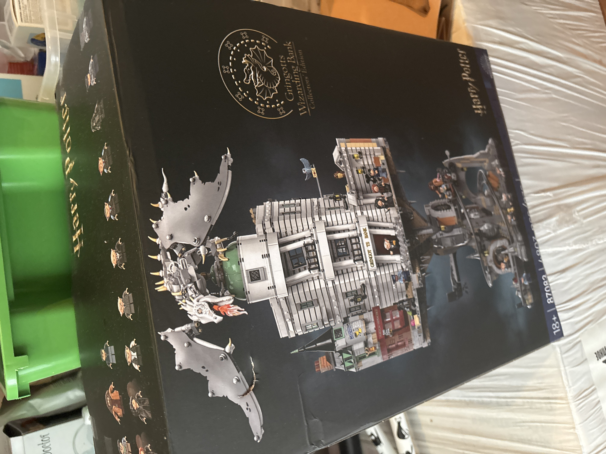 The image shows a black box, most likely a product packaging, featuring a depiction of Gringotts Wizarding Bank from the Harry Potter series. The box is angled slightly and is surrounded by other objects, indicating it is likely indoors. The box has images of the bank building, a dragon, and several minifigures. The words "Gringotts Wizarding Bank Collectors Edition" and "Harry Potter" are prominently displayed on the box. A small "+18" and a string of numbers can also be seen on the lower right corner of the box.