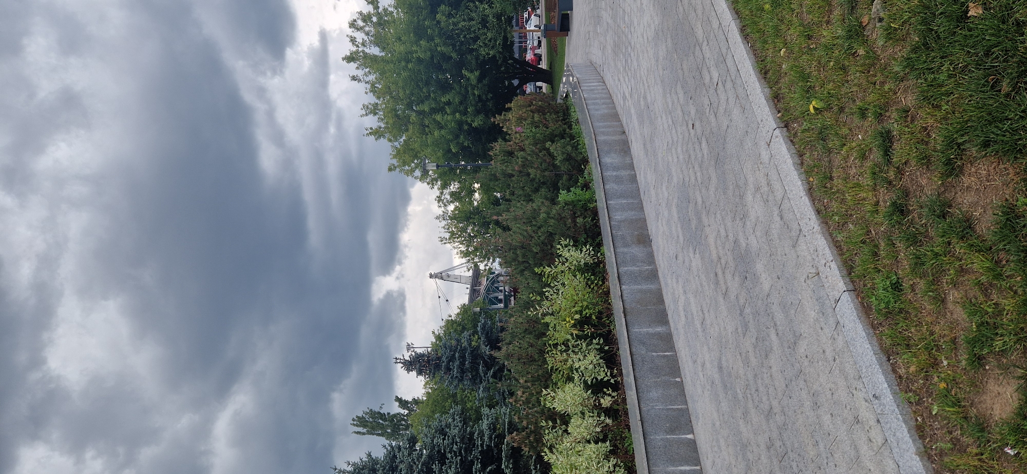 The image depicts an outdoor scene with a paved walkway in the foreground and a landscaped area with trees and shrubs behind it. The sky is overcast with dark clouds. In the background, a large industrial structure resembling a crane or tower is visible amongst the trees. To the right, a portion of a building with cars parked in front of it can be seen. The scene appears to be a public park or a similar urban outdoor space. The location is not specified in the image itself, and therefore cannot be identified as Moscow, Russia.