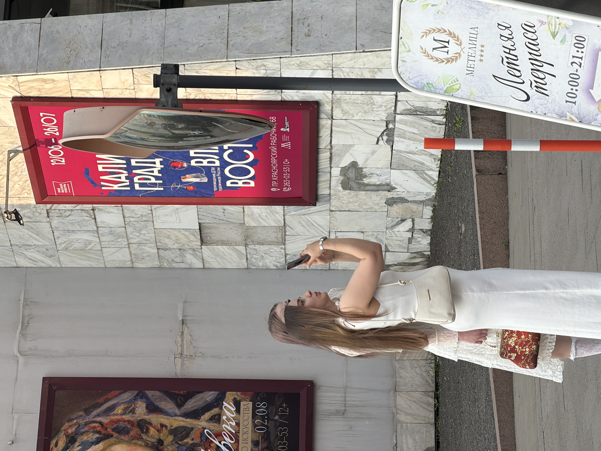 The image captures a street scene in Krasnoyarsk, Russia, featuring two young women in white dresses standing near a building with a marble facade. One woman is taking a photo with her phone. A large red advertising board with Russian text and a convex mirror is prominently displayed on the wall. A white A-frame sign with the inscription "МЕТЕЛИЦА" and "Летняя терраса" is visible in the foreground, suggesting a nearby establishment. The lighting indicates it is daytime, likely late afternoon due to the shadows.