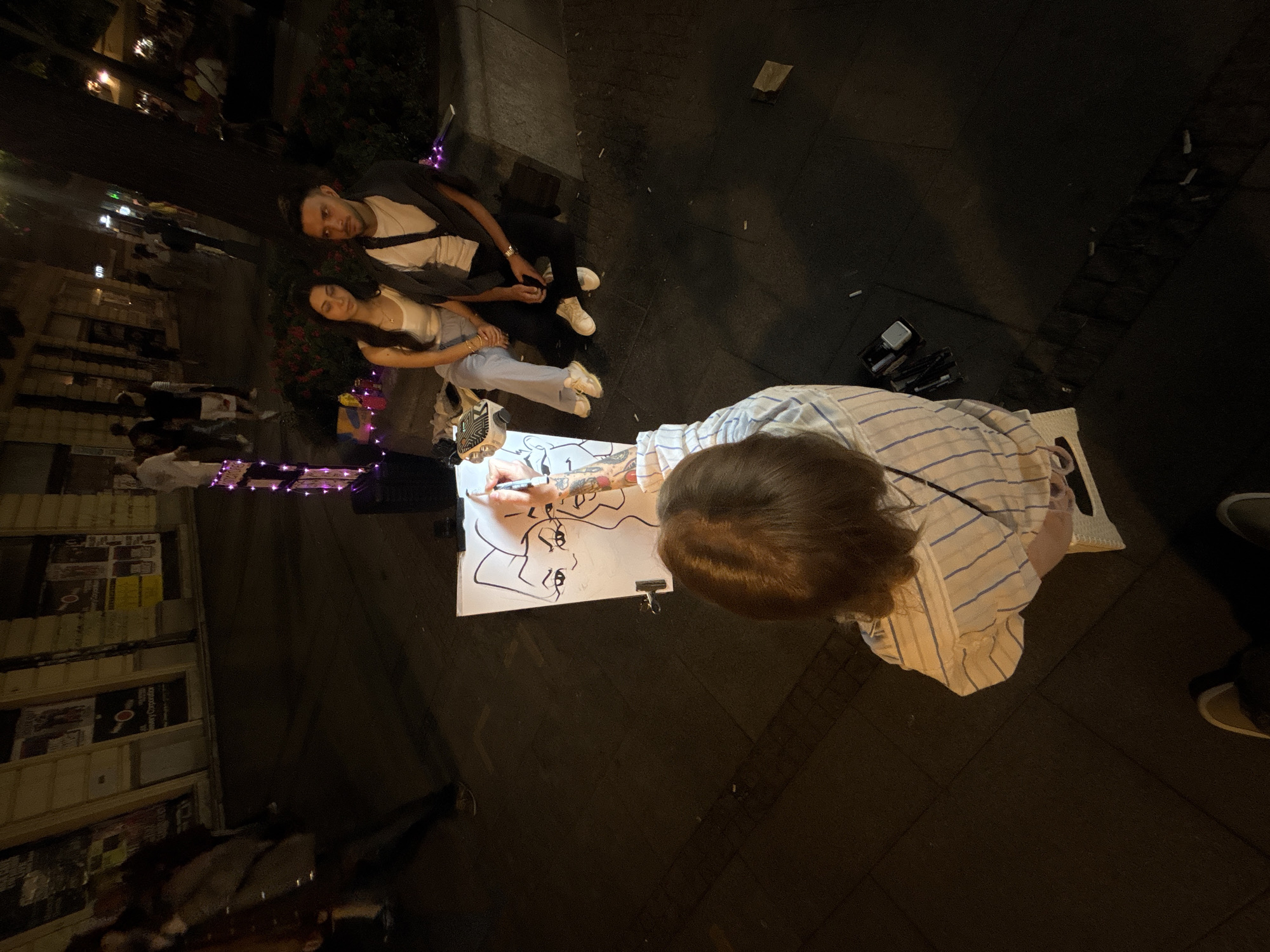 An eye-level, high-angle outdoor shot captures a street scene at night, likely in Belgrade, Serbia, with a focus on an artist drawing a caricature. The foreground shows the back of a person, presumably the artist, wearing a striped shirt, seated and drawing on a white easel. The drawing on the easel features exaggerated facial features of two people. To the right of the artist, a young couple is seated on a low wall or bench, appearing to be the subjects of the caricature. The woman wears a white tank top and light blue pants, while the man wears a white t-shirt, dark pants, and a dark open cardigan. They are looking towards the artist.
The background is a dimly lit street with shop fronts. Some people are visible walking in the distance, and a tree with red flowers is to the right of the couple. The street is paved with dark tiles. A small stand with purple lights is also present, possibly for display or illumination. The overall atmosphere is that of a lively urban evening, with the focus on a local artist at work.