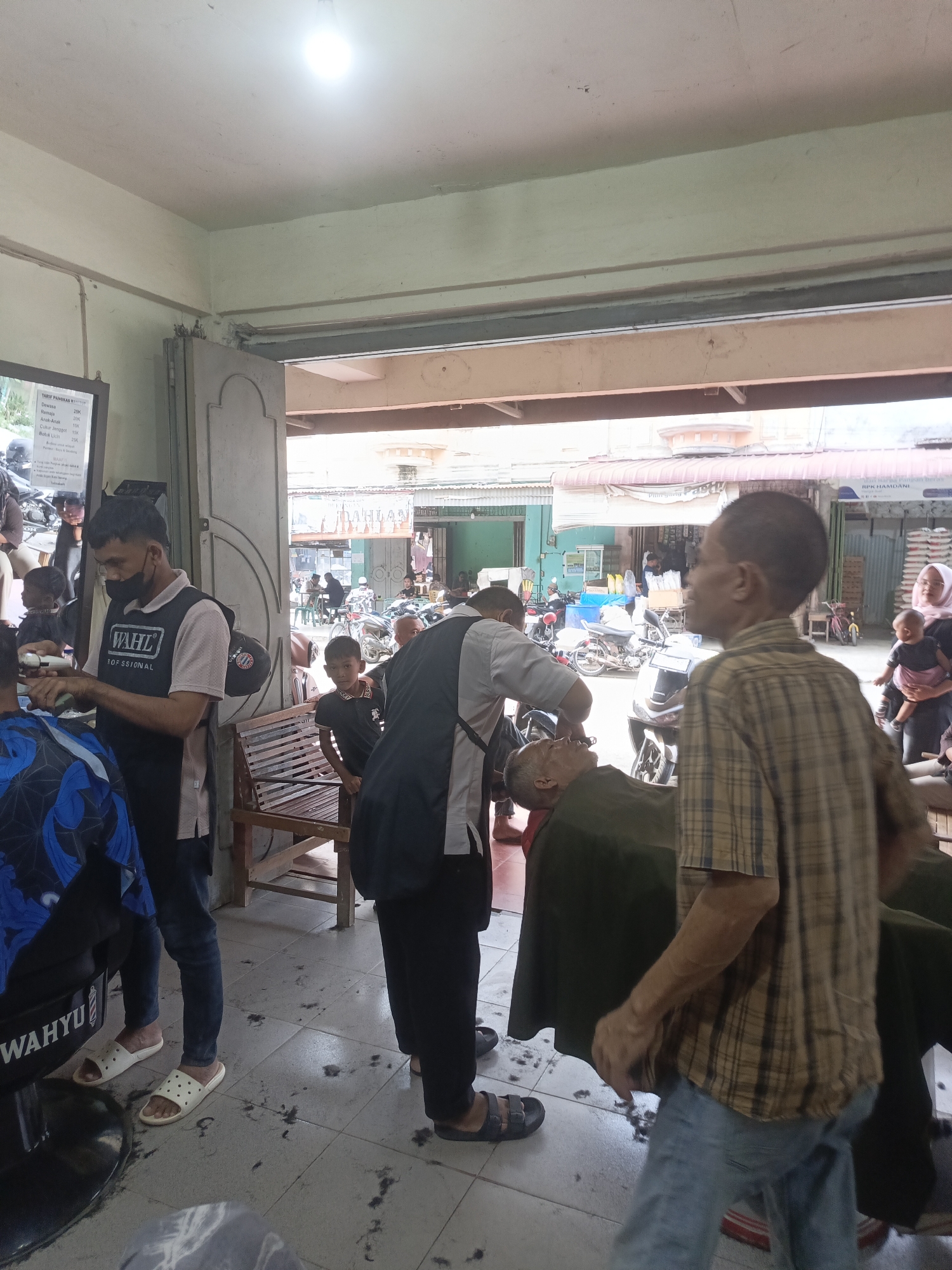 The image depicts a barbershop in Lhokseumawe, Indonesia. Inside the shop, a barber, wearing a mask and an apron with "WAHL PROFESSIONAL" printed on it, is in the process of cutting the hair of an elderly man who is seated in a barber chair, draped with a dark green cloth. Another man in a checkered shirt and jeans is walking past the barber chair, looking towards the right side of the frame. In the background, several other people are visible, including children and a woman holding a baby. The shop appears to be open to the street, with shops and motorbikes visible outside. The floor of the barbershop is littered with cut hair. A price list for haircuts is also visible on the wall.