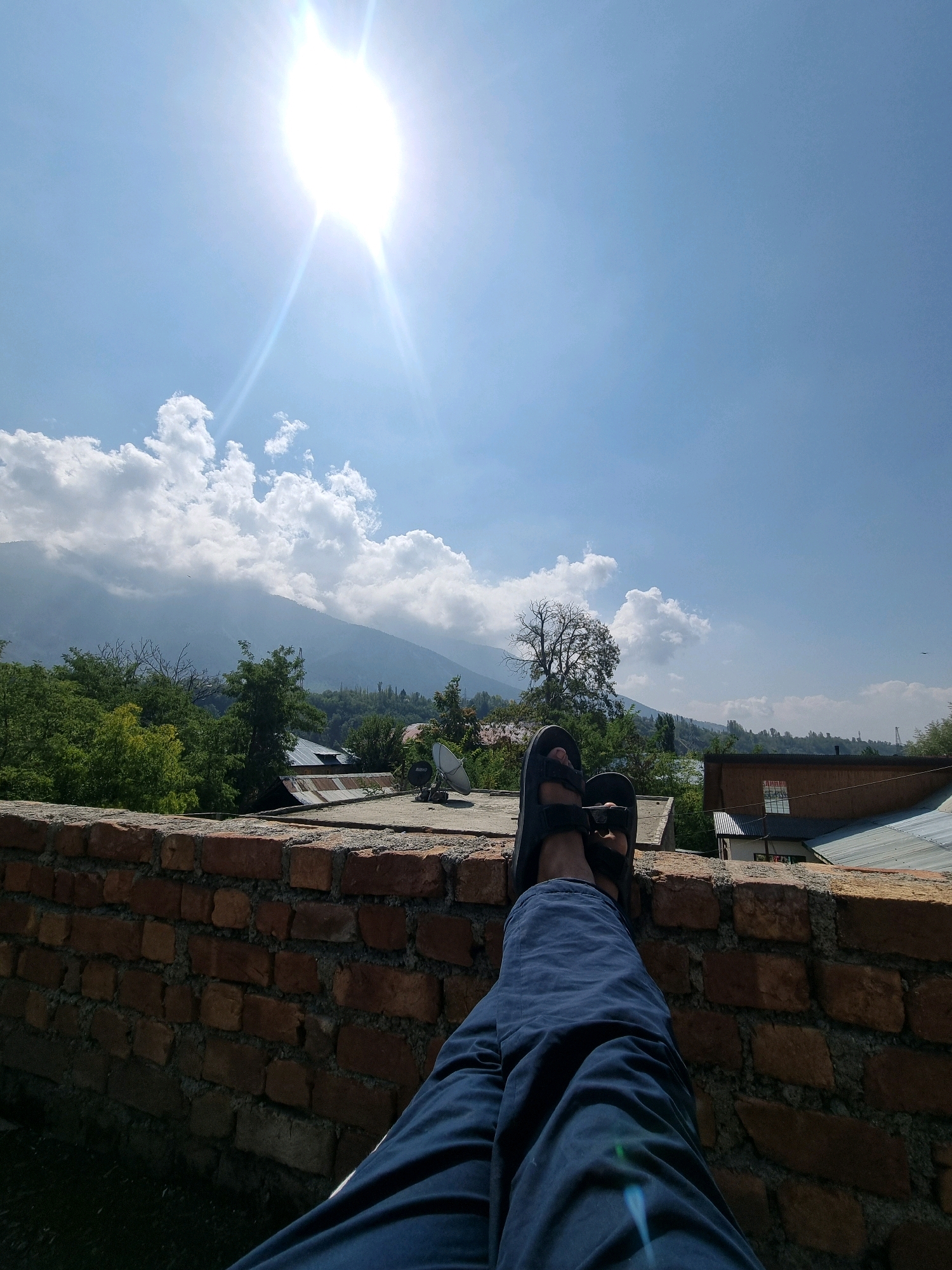 The image depicts a person's legs and feet, clad in blue pants and dark sandals, resting on a brick wall in Manigam, India. The wall is constructed from reddish-brown bricks with visible mortar. Behind the wall, rooftops of buildings are visible, some with metal sheeting. Lush green trees and vegetation are scattered throughout the landscape. In the background, a large mountain range is shrouded in mist, with a bright sun shining in the clear blue sky, casting lens flares. Scattered white clouds are present in the sky. The scene appears to be outdoors, possibly from a rooftop, offering a view of the surrounding environment under a sunny sky.