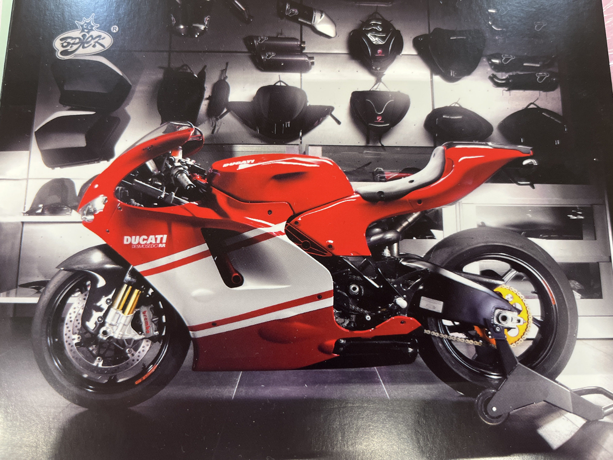The image shows a red and white Ducati motorcycle. The motorcycle is positioned vertically in the frame, with its front wheel at the top. The motorcycle is on a stand, with its rear wheel elevated. The background appears to be a workshop or showroom, with shelves filled with helmets and other motorcycle accessories. The lighting is bright, and the image is sharp.
The location is not specified in the image or provided in the text. The image does not provide any information about the city of Kharkiv, Ukraine.