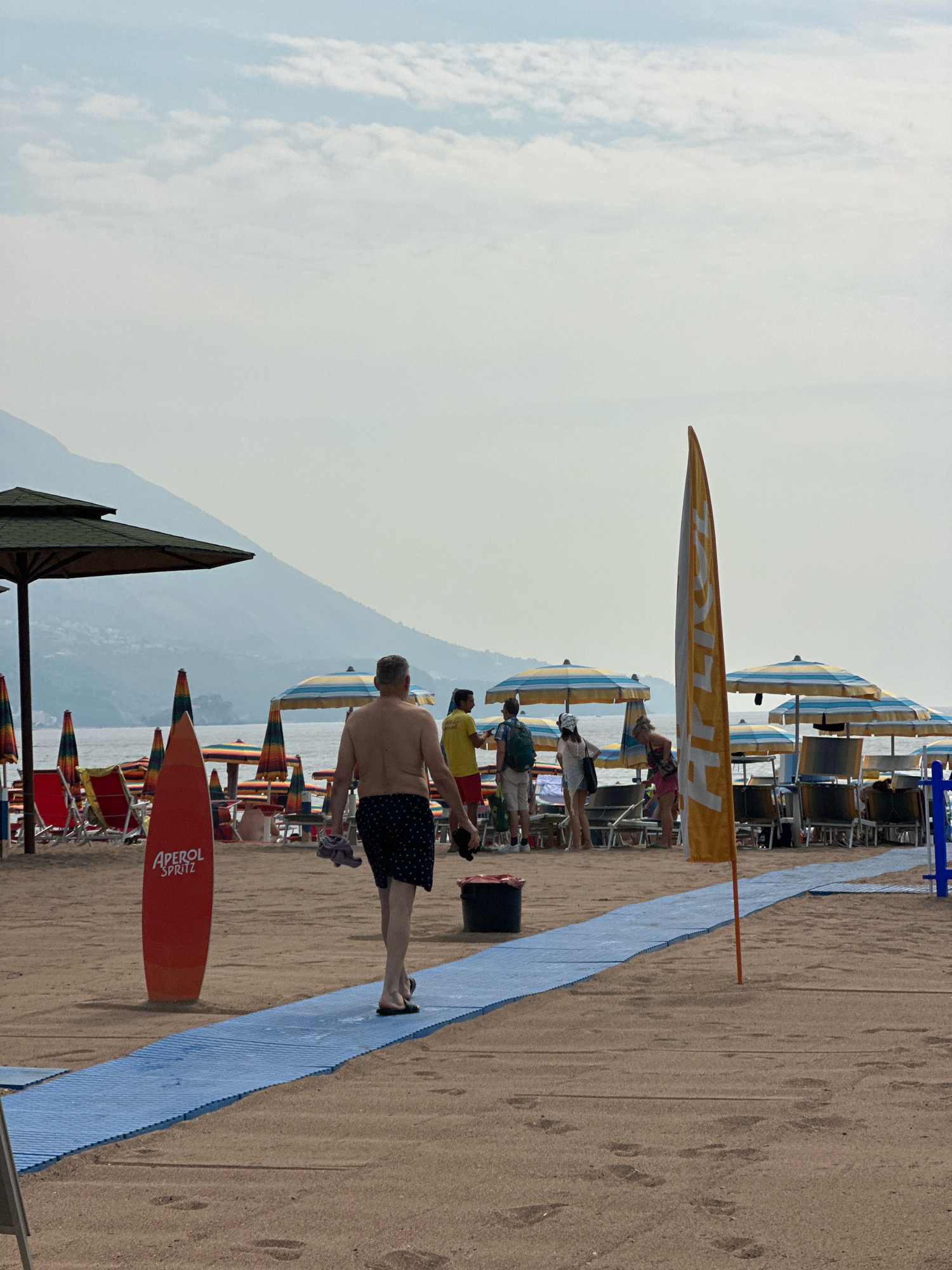 The image depicts a sunny day at a beach in Boreti, Montenegro. A man, shirtless and wearing patterned shorts, walks away from the camera on a blue mat laid across the sandy beach. He is holding a dark object in his right hand and a light colored towel in his left. To his left, a tall orange surfboard-shaped sign with "APEROL SPRITZ" written on it is planted in the sand. In the background, several beach umbrellas with yellow, blue, and white stripes are set up, with some chairs visible underneath. Other people are milling around the beach, some standing and others seated. In the distance, mountains form a hazy backdrop against a pale sky with scattered clouds. The sea is visible between the beach umbrellas and the mountains.