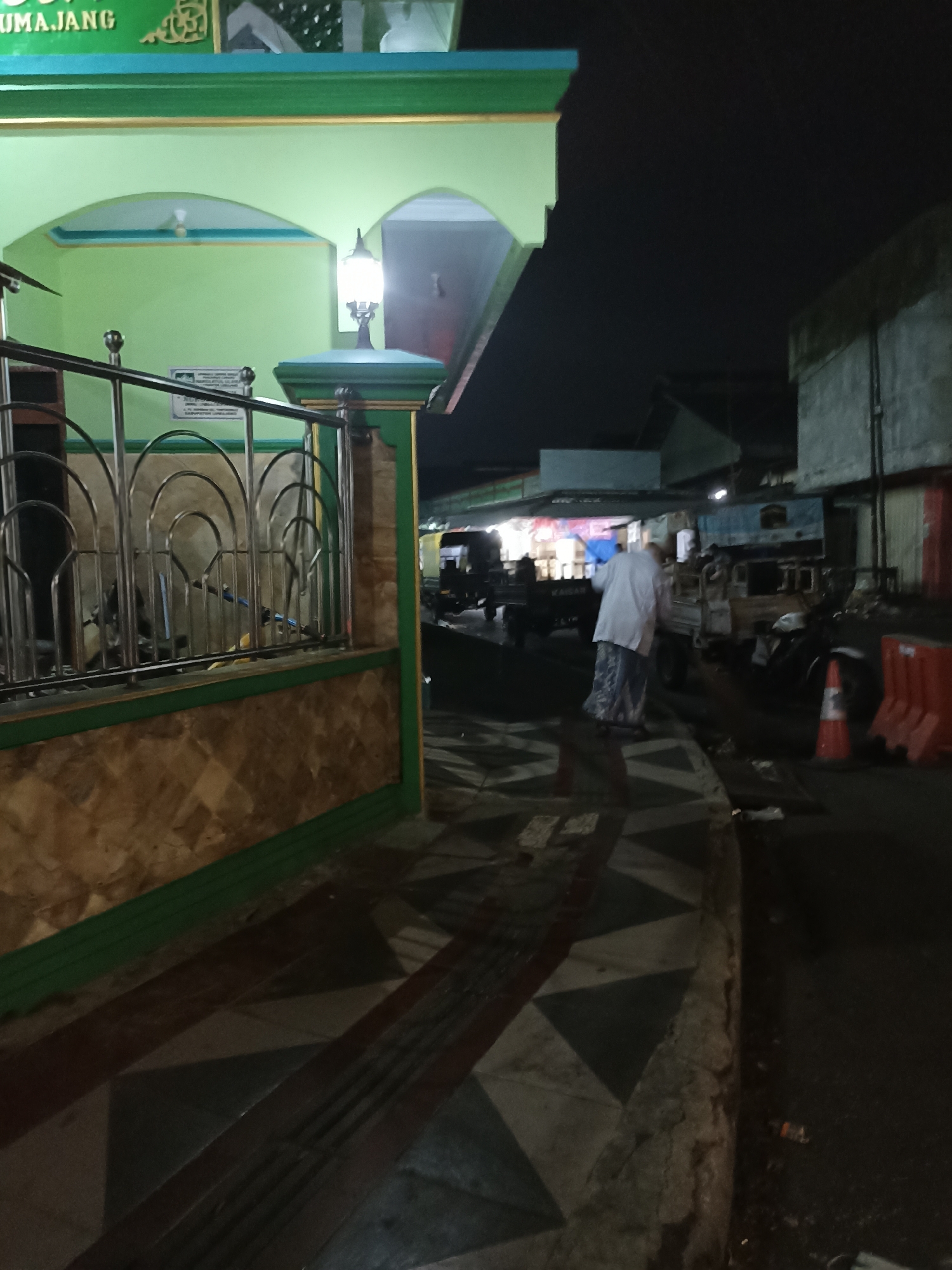 The image shows a nighttime street scene in front of a building with a green facade and a patterned wall. The building appears to be a mosque, with the word "TUMAJANG" visible on an upper section. A metal railing with decorative arches is in the foreground. A person wearing a white long-sleeved shirt and a patterned sarong is walking away from the camera, pushing a broom. Several vehicles, including a motorcycle and what appear to be small trucks, are parked on the street behind the person. There are also orange traffic cones and what looks like debris on the ground. The lighting is dim, with a streetlamp on the building illuminating part of the scene. The street has a patterned tiled walkway in the foreground. The overall impression is a quiet street, possibly near a place of worship, at night.