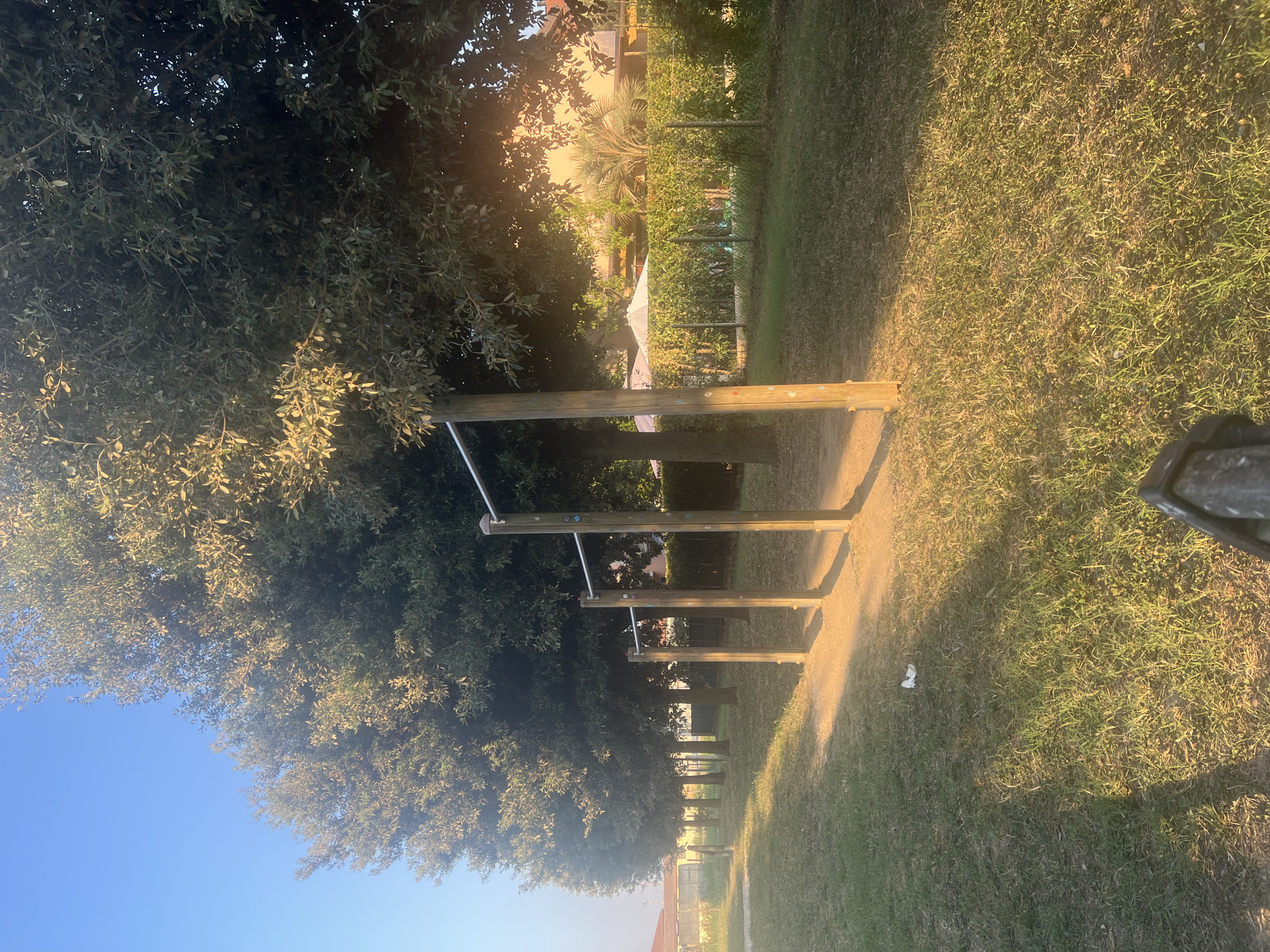 The image depicts an outdoor scene with rows of trees casting shadows on a grassy area. In the foreground, there are several horizontal bars supported by wooden posts, resembling an outdoor fitness equipment setup. The ground is covered with grass, and there is a small piece of white debris on the ground. In the background, there are more trees, a hedge, a fence, and some buildings with umbrellas visible. The lighting suggests it is either early morning or late afternoon, with the sun casting long shadows. The location is identified as Sant'Eufemia, Italy.