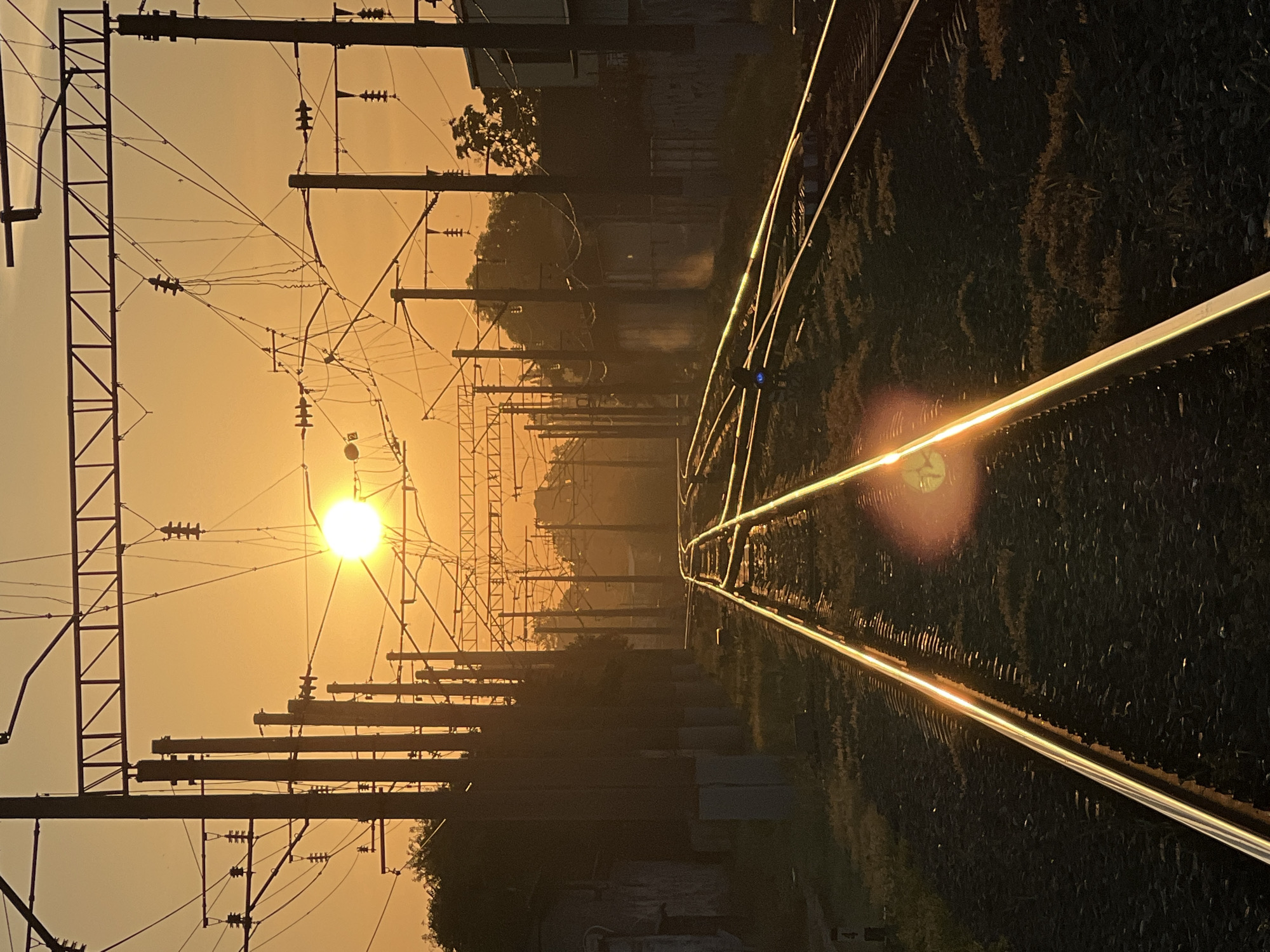 The image depicts a railway track in Krasnodar, Russia, during sunset. The sun is low in the sky, casting a warm orange glow across the scene. The railway tracks lead into the distance, disappearing into the hazy horizon. Overhead, a complex network of power lines and supporting poles crisscrosses the sky, silhouetted against the bright sunset. Trees and buildings are visible on either side of the tracks, also silhouetted. The foreground of the image focuses on the gleaming metal of the railway tracks, reflecting the sunlight. The ballast of the track is visible, with some sparse vegetation growing between the stones. The overall atmosphere is one of quiet solitude and the end of the day.