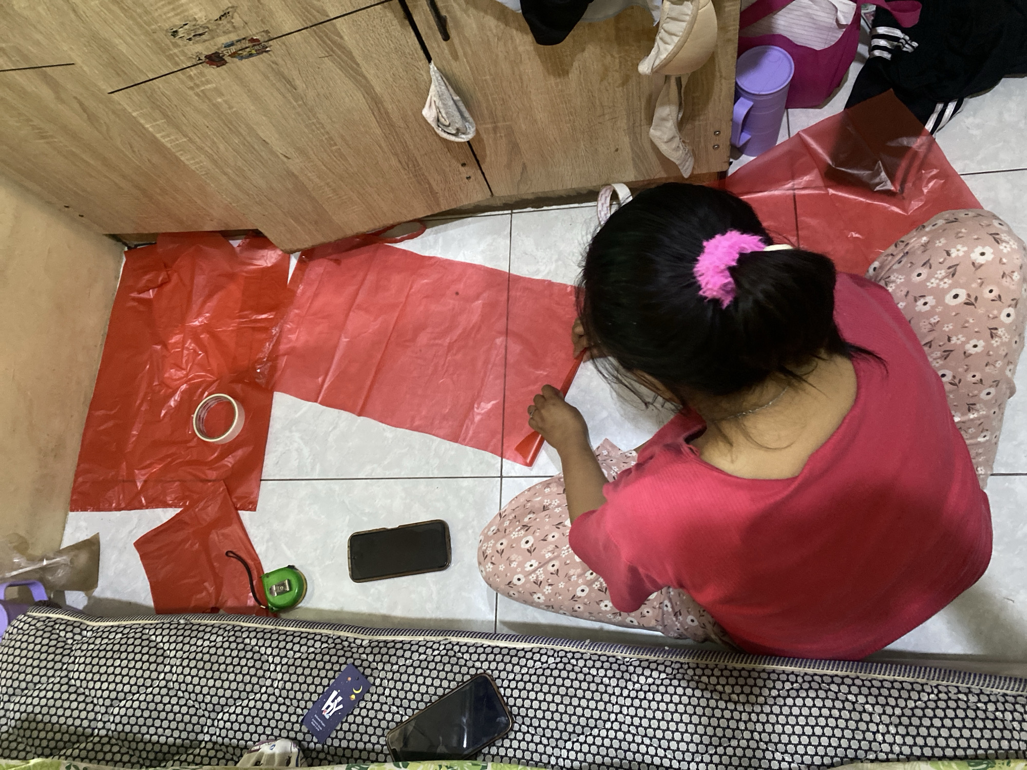 A person, viewed from above, sits on the floor amidst scattered items. The person is wearing a pink top and patterned pants. Red plastic sheeting is spread out on the floor, with a roll of tape and a measuring tape nearby. Two smartphones are visible on the floor, one to the left of the person and another further up. To the left of the scene is a mattress or bed with a black and white patterned cover. To the right, a wooden cabinet is partially visible, with some folded clothing and purple containers on the floor beneath it. The setting appears to be a residential interior, possibly in Cilegon, Indonesia, given the context.