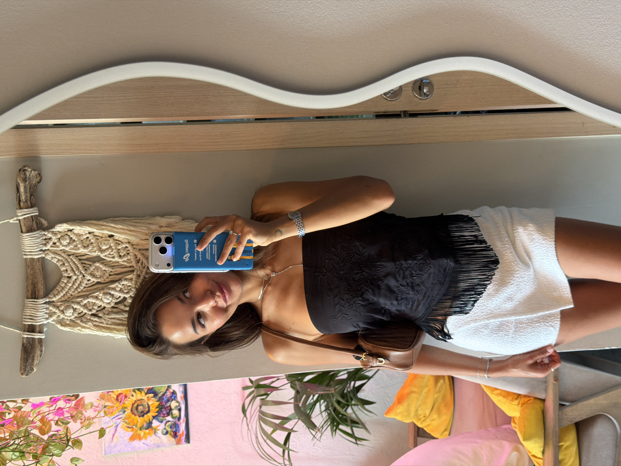 A young woman is taking a selfie in a mirror in an indoor space. She is wearing a black strapless top with fringe detailing and a white textured skirt. She has a brown purse slung over her shoulder. The background includes a macrame wall hanging, a painting of sunflowers and fruit, and a potted plant. A large mirror is on the right side of the frame, with a wooden door frame visible behind it. The overall environment appears to be a stylish room with bohemian decor. The location is specified as Sochi, Russia.