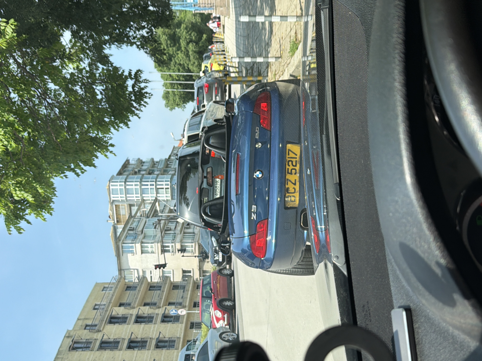 The image, oriented vertically (rotated 90 degrees counter-clockwise from typical landscape orientation), captures a bright daytime urban street scene in Varna, Bulgaria, viewed from inside a vehicle looking out the front passenger window.
In the mid-ground, a blue BMW Z3 roadster is prominently featured, facing away from the viewer. The car's model "Z3" and "2.3" engine designation are visible on its rear. Its Bulgarian license plate is yellow with black characters, reading "BCZ 5217". Directly behind the BMW is a dark-colored SUV, with a partially visible Bulgarian license plate "4056 HK". Further down the street, a red Mini Cooper and other vehicles are part of a traffic queue.
The street is lined with multi-story buildings of varying architectural styles. On the left, a building features distinctive tiered balconies or exterior fire escapes. Another taller structure in the background boasts a modern upper section with extensive glazing. Lush green trees are visible along the street, complementing the urban landscape.
A sidewalk runs alongside the road, delineated by a row of black and white striped bollards. A traffic light and a circular blue traffic sign are also visible in the distance. The sky is clear and bright blue, indicating sunny weather. The overall scene depicts a typical busy city street under clear conditions.