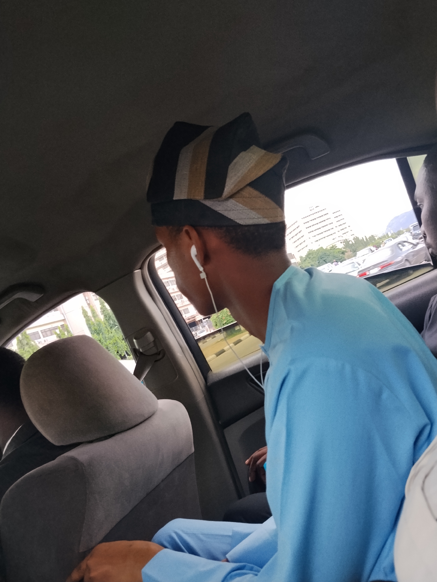 The media file shows a medium shot from inside a car, looking towards the passenger side. The primary subject is a young Black man wearing a light blue dashiki and a traditional striped Aso Oke cap. He has white earbuds in and is looking out the window. The car's interior is visible, with grey fabric seats and the headliner. Through the window, there are buildings, trees, a body of water, and other cars on what appears to be a road. The lighting suggests it is daytime, with bright, overcast conditions. The scene appears to be in an urban environment in Abuja, Nigeria, possibly near a waterfront area given the water visible. The man's focus suggests he is observing the outside world, and there are no strong emotional indicators conveyed. No visible text is present in the image.