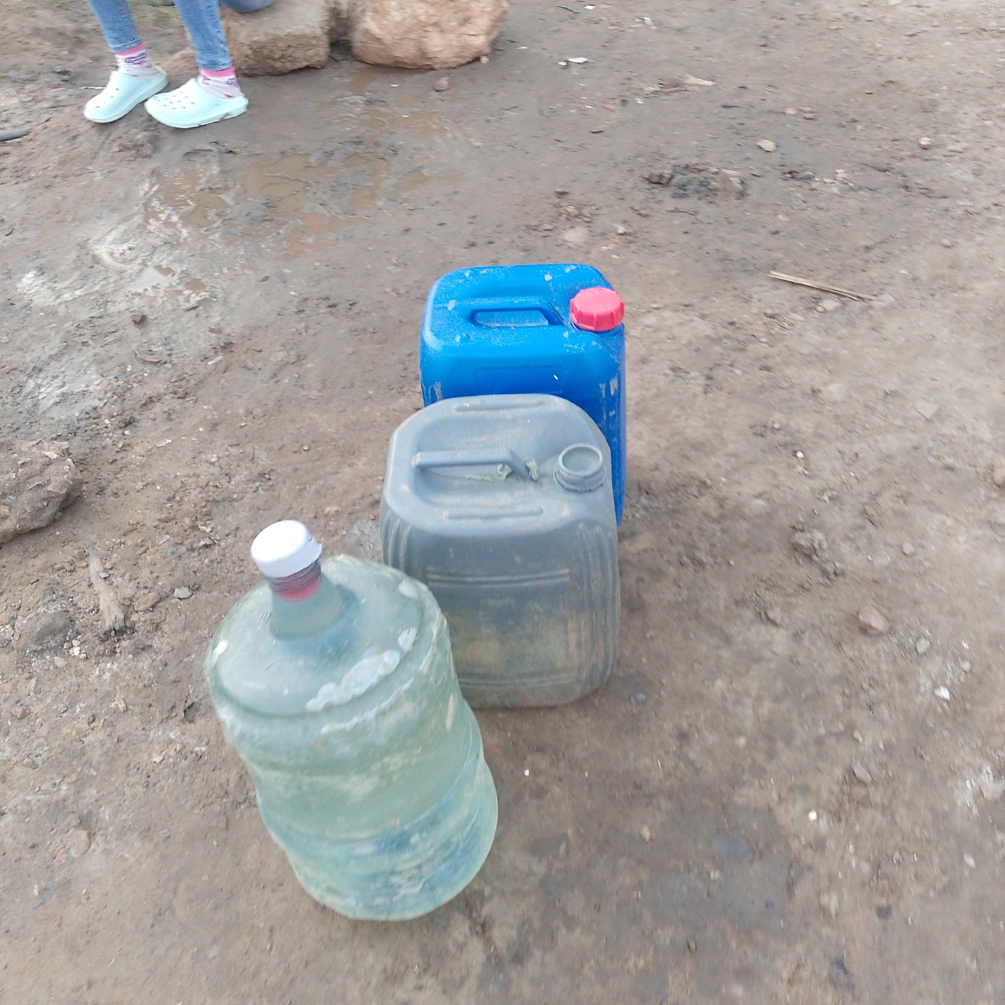 The image shows three containers of water or other liquids sitting on a muddy dirt ground. One is a blue rectangular container with a red cap, another is a gray rectangular container, and the third is a large round plastic bottle with a white cap. In the top left corner of the image, there are legs of a person wearing light blue shoes and socks. The location is possibly in Altagracia, Venezuela, given the user's request. The ground is uneven and appears to be dirt mixed with mud or wet soil.