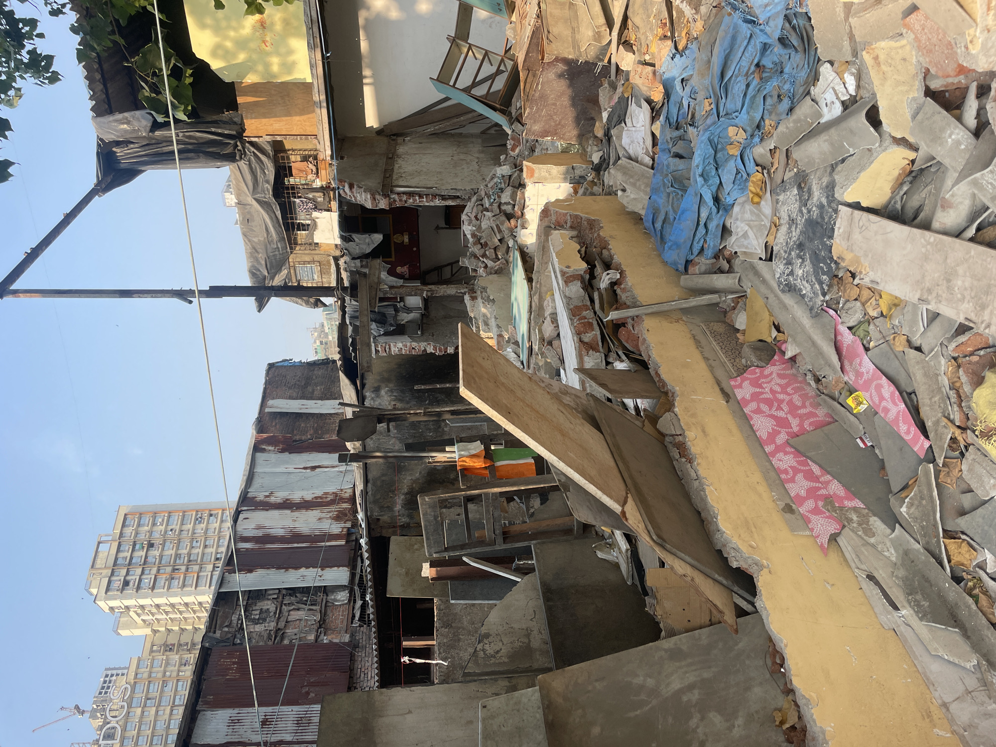 The image shows the aftermath of a building collapse in Mumbai, India. Debris litters the scene, including broken bricks, concrete slabs, and personal belongings. A neighboring high-rise building is visible in the background, contrasting with the dilapidated conditions of the collapsed structure. The scene suggests significant damage and potential loss of property and life.