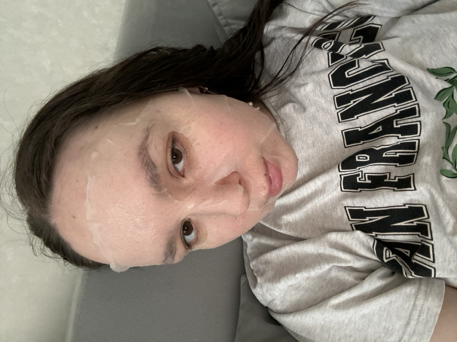 A female individual is depicted in a close-up, head-and-shoulders shot, in an indoor setting within the city of Chaikovsky, Russia. She has brown hair and brown eyes, and her face is covered by a translucent white sheet mask, which appears moist with visible liquid or serum. The mask features cutouts for her eyes, nostrils, and mouth. She wears a light grey, short-sleeved t-shirt with dark lettering on the front, partially legible as "SAN FRANC...", likely part of "SAN FRANCISCO". A small, light-colored, possibly pearl-like, earring is visible in her right ear. Her gaze is directed towards the camera, and her lips are slightly parted. The background consists of a plain, light-colored wall in the upper left and a grey upholstered surface behind her right side, indicating she is seated or reclining.