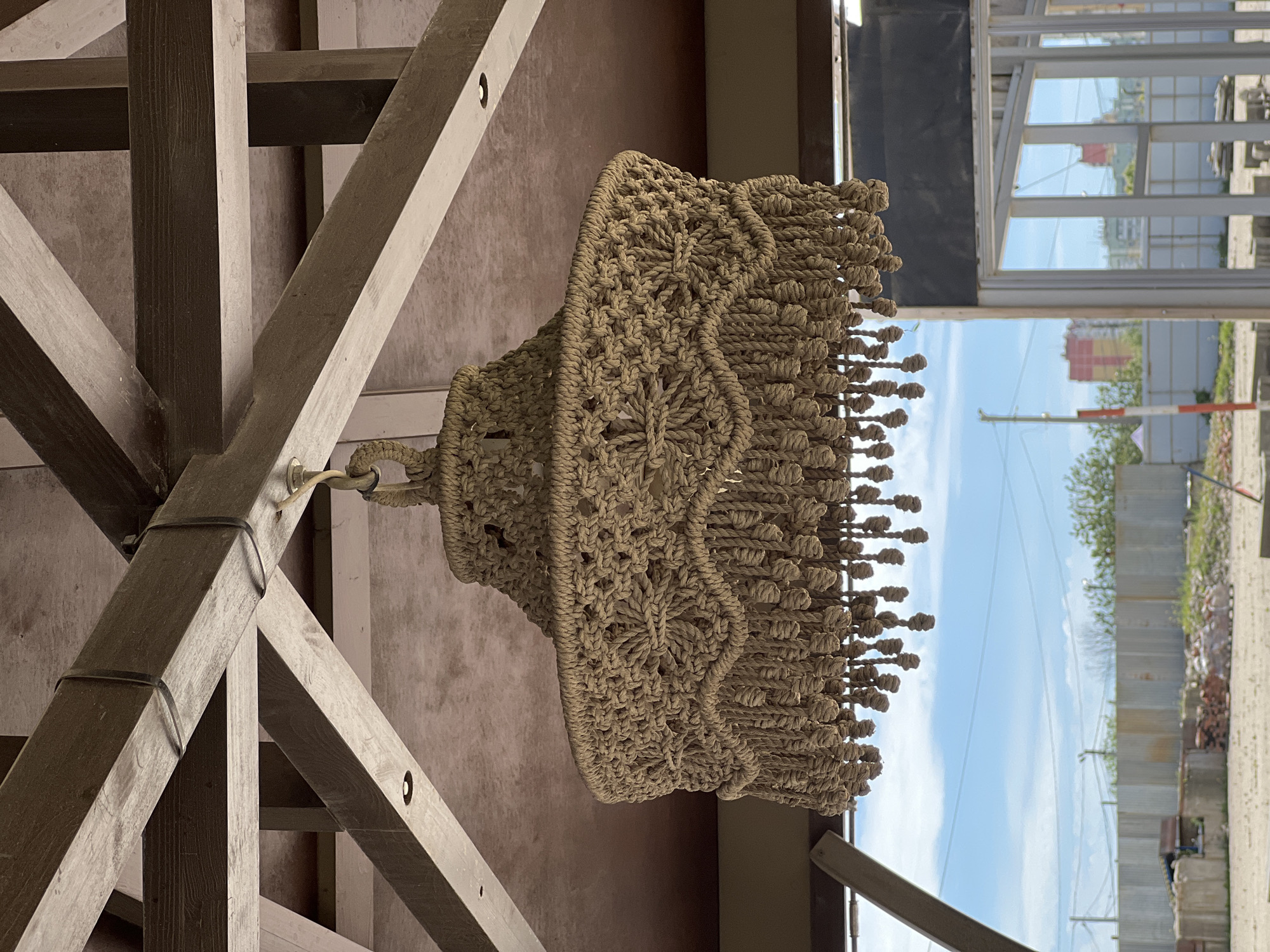 The image is rotated 90 degrees clockwise, depicting a macrame light fixture hanging from a wooden ceiling structure, with an outdoor scene visible through a large opening to the right.
The central subject is a large, beige or light brown macrame chandelier or decorative lampshade. It is intricately hand-knotted from thick rope or cord, featuring a tiered design with elaborate patterns, including diamond shapes and floral motifs created through various macrame knots. The bottom edge of the fixture is adorned with numerous short, knotted fringes, forming a decorative border. The macrame piece is suspended by a braided rope loop, attached to a metal fixture on one of the wooden beams.
The fixture hangs beneath a robust overhead wooden beam structure, which dominates the upper left and central portions of the image. These beams vary in shade from dark to light brown, displaying visible wood grain and suggesting a rustic or natural aesthetic. They are arranged in an interlocking or criss-cross pattern, indicative of a ceiling or pergola support. Some beams show minor wear, small holes, or metal connectors, and one section has black bands wrapped around it, possibly for structural integrity or utility.
To the right of the macrame and wooden structure, a large window or opening reveals a bright, clear blue sky with some scattered white clouds. In the distance, parts of buildings, including one with a red roof, and patches of green foliage are visible, suggesting an urban or semi-rural landscape in Krasnodar, Russia. A light-colored barrier, resembling a road or construction barrier, can be seen in the lower right portion of the outdoor view.
The scene takes place during the daytime, characterized by abundant natural light and a clear sky. No people are visible in the image, and there is no discernible text. The overall atmosphere is tranquil, showcasing craftsmanship and an open environment.