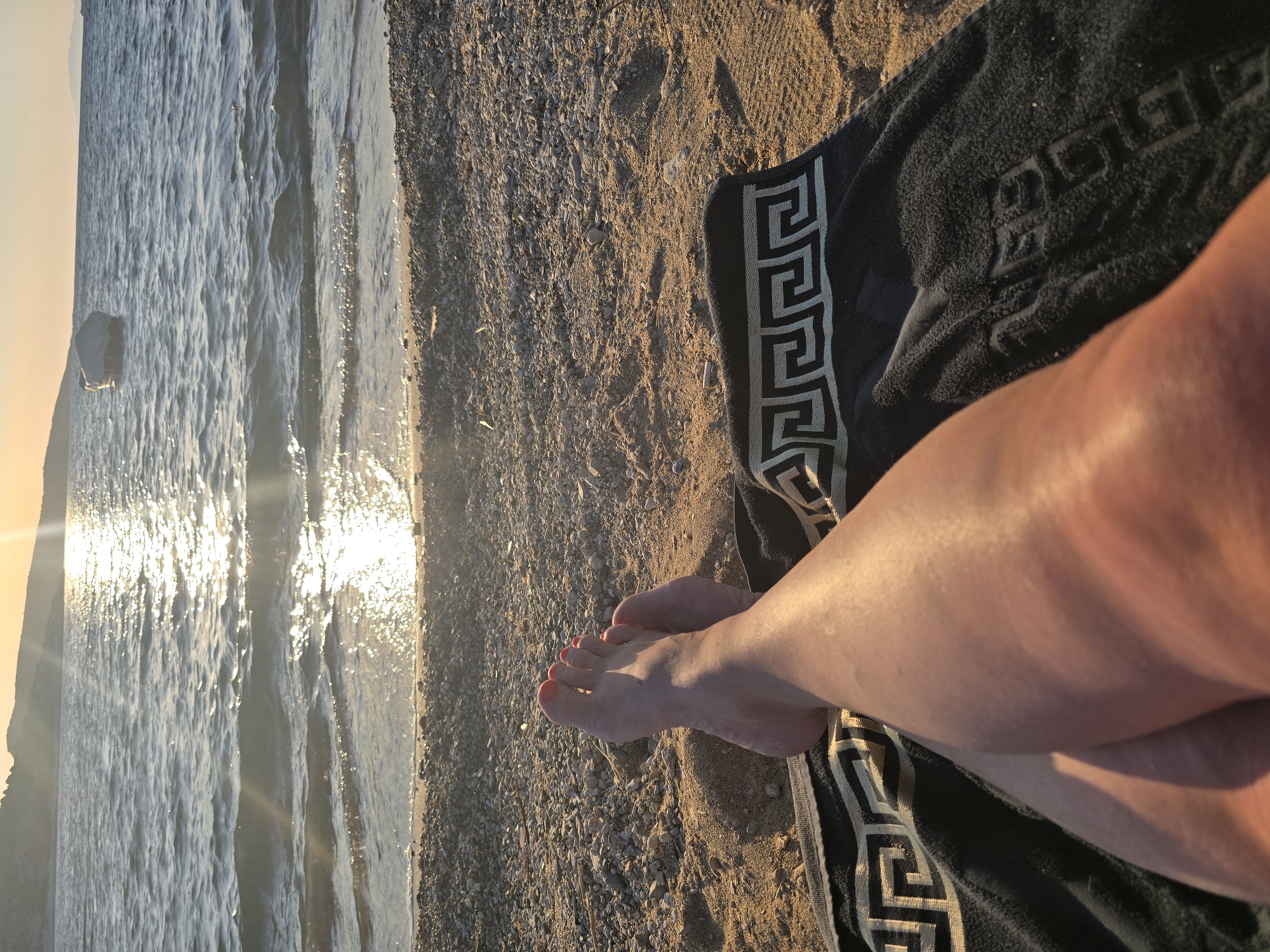 A first-person perspective shot captures a person's legs and feet resting on a beach in Foça, Turkey. The individual is lying on a black towel with a Greek key pattern, the lower half of their legs and feet in the foreground. Their feet are bare, with red nail polish on the toenails. The beach itself is covered in sand and pebbles, with the gentle waves of the sea in the background. The sun is setting or rising, casting a warm, golden glow across the water and the scene, creating sun flares and reflecting brightly on the sea's surface. A small boat is visible in the distance on the water. The overall impression is one of relaxation and tranquility at the seaside.