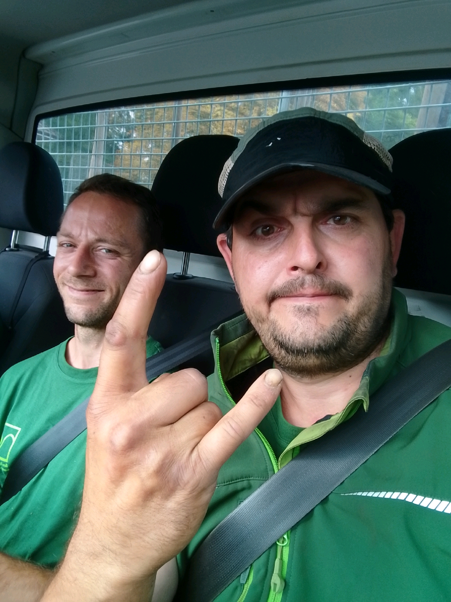 The image shows two adult males in the interior of a vehicle, likely a van, as suggested by the safety grates behind them. The man in the foreground, on the right, is wearing a black and green baseball cap and a green jacket with a zipper. He is making a "rock on" hand gesture with his right hand and is looking directly at the camera. The man behind him, on the left, is wearing a green t-shirt and is smiling and looking slightly to his left. Both men appear to be in good health. The interior of the vehicle is visible, including black seats and a gray seatbelt across the man in the foreground. Outside the vehicle, through the rear window, a blurred background of trees and foliage is visible, suggesting a suburban or rural setting. The image is taken from a front-facing perspective, likely by one of the occupants. There is no clear indication of the specific location within Wedel, Germany, but the presence of trees and foliage suggests it is not in a densely populated urban area.