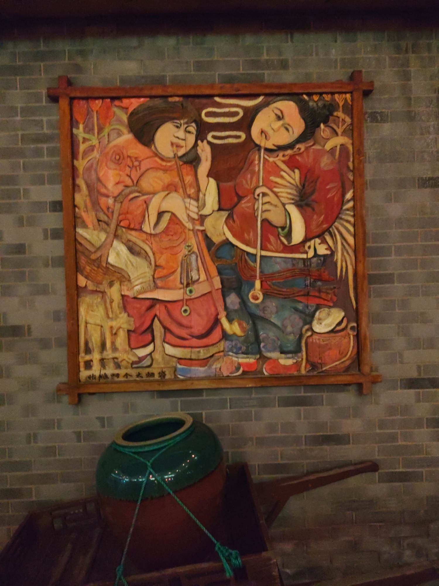 This is a medium shot of a decorative wooden carving depicting two figures, possibly from ancient China, in an indoor setting against a brick wall. The carving is framed by dark wood. The left figure has dark hair pulled back, a round face with rosy cheeks, and is wearing an orange robe with patterned sleeves and a pink skirt. This figure holds a thin pipe to their mouth. The right figure also has dark hair, a more angular face, and is wearing a red robe with white cuffs and a patterned blue skirt. This figure is also holding a thin pipe to their mouth. Both figures appear to be engaged in the activity of smoking. The background of the carving is rendered with red and yellow floral motifs. Below the carving, a large, dark brown wooden cart holds a large, round, dark green ceramic pot with a rim. Green rope is tied around the pot, with some of the rope extending to the cart. The cart has a long, dark wooden handle. The wall behind the carving and the pot is made of grey bricks. The lighting appears to be artificial and slightly dim. This scene is located in Lindian, China.