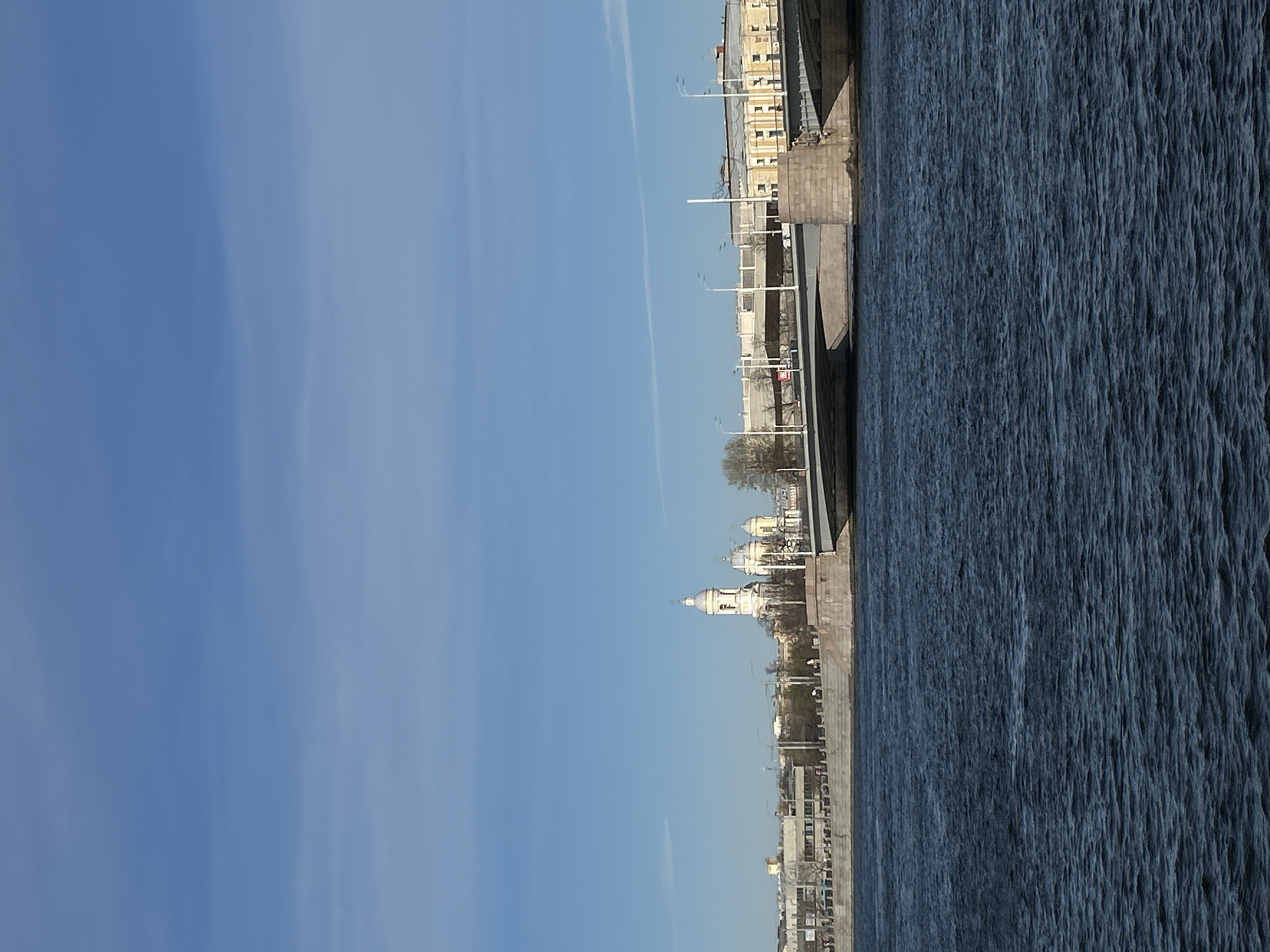 The image captures a cityscape in Saint Petersburg, Russia, featuring a wide expanse of water along the right side and a clear, light blue sky on the left. Buildings line the embankment; the architecture features light-colored facades, and there are some architectural details such as domes, seen in a distance. The water surface has texture, suggesting it is not completely still. Streetlights are visible along the embankment. The sky is clear, with what appears to be condensation trails from an aircraft running across it.