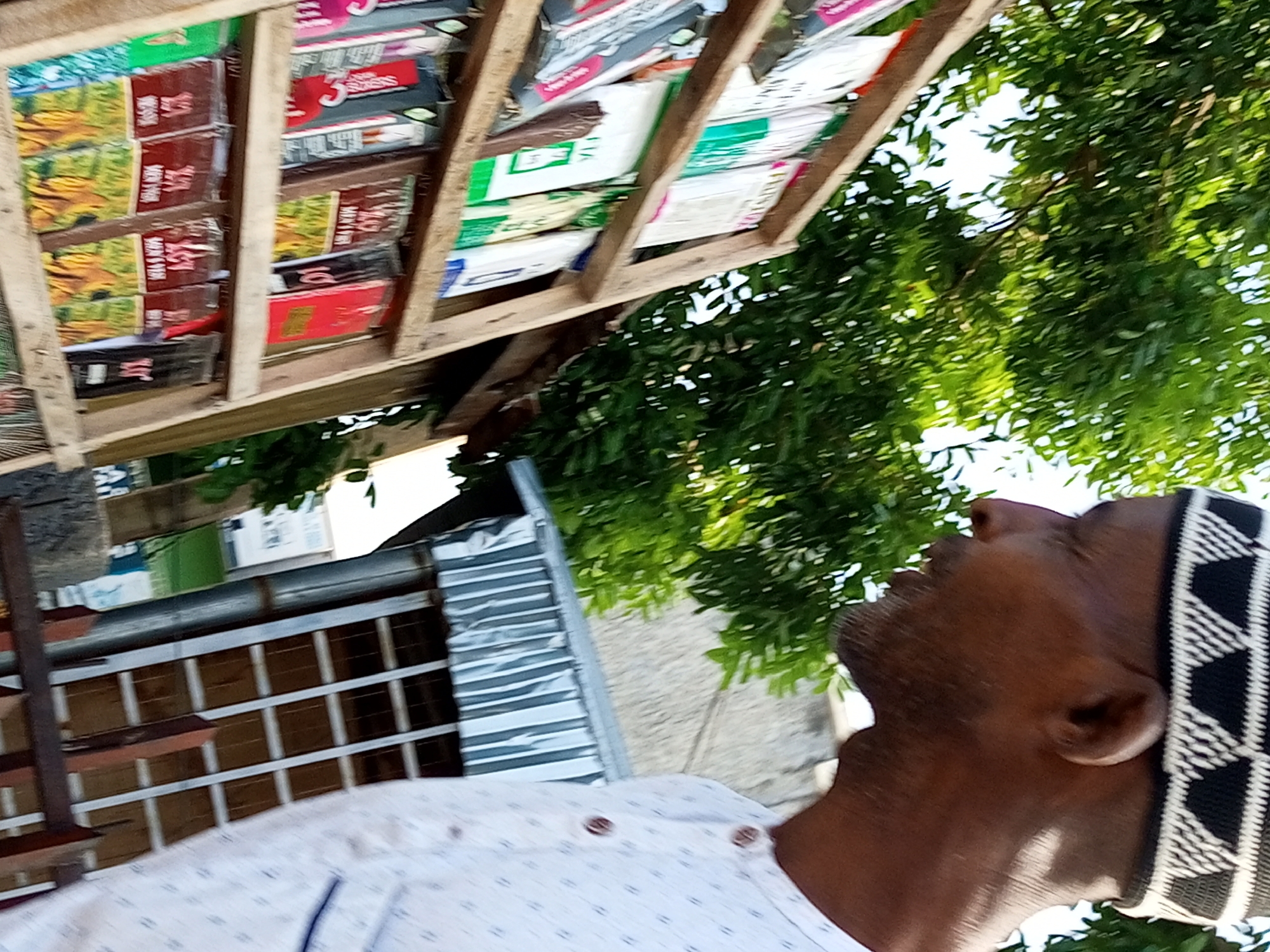 In Tempure, Nigeria, an overhead shot captures a person wearing a white shirt with a subtle dotted pattern and a black and white kufi hat. The individual is looking upwards. The scene is set outdoors, with lush green trees overhead. In the foreground, a wooden shelf unit is filled with various boxed items, possibly goods for sale. Below the shelf, a metal grating structure and a portion of a building are visible. The image is taken from a low angle, looking up towards the sky, with the shelves and the person forming diagonal lines in the composition.