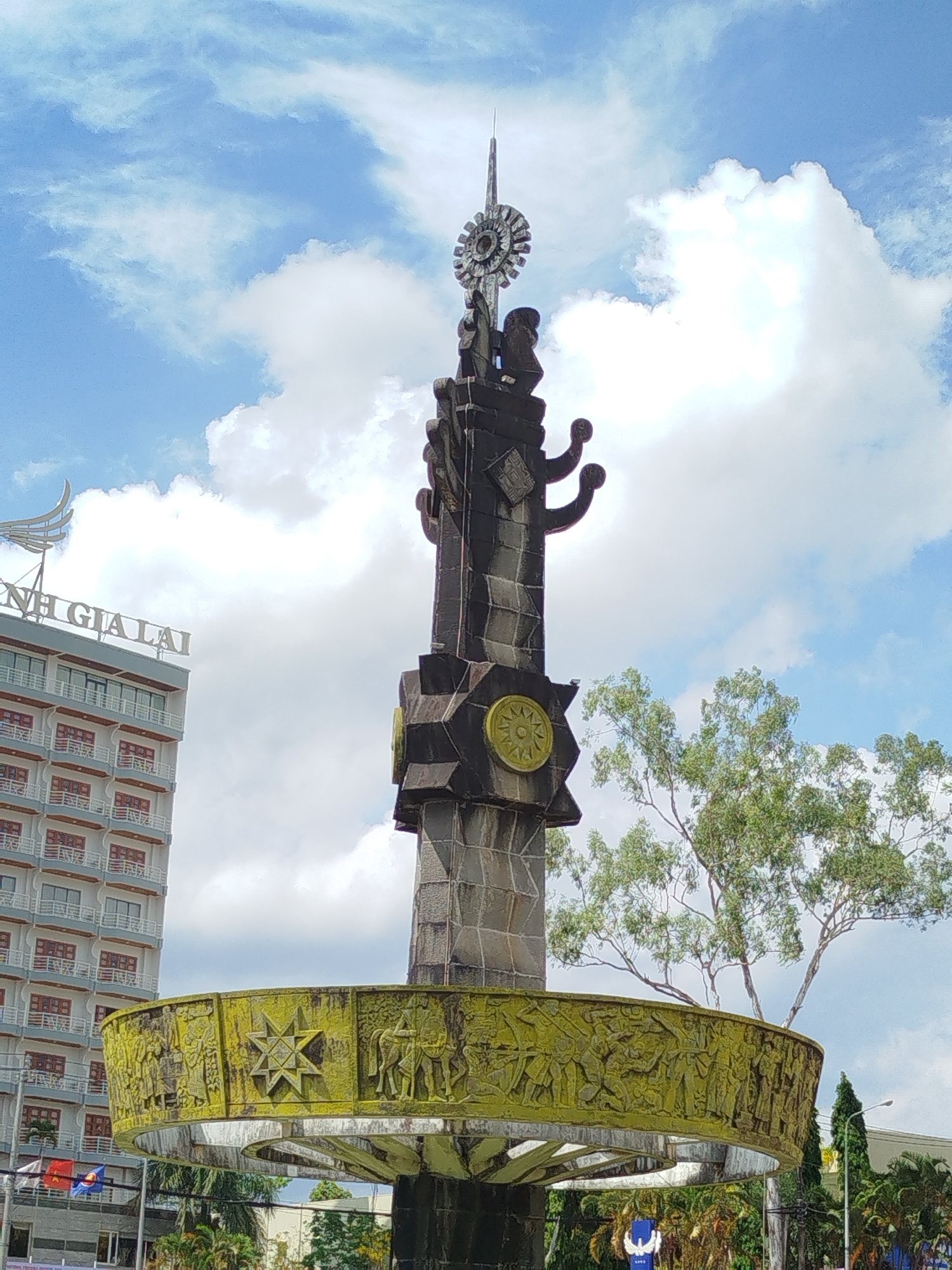 The image depicts a tall, sculpted monument in Pleiku Ward, Vietnam, under a partly cloudy sky. The monument features a tiered base with intricate carvings, a central shaft adorned with geometric and circular motifs, and a sunburst-like emblem at the top. A wide, circular element with additional carvings encircles the lower portion of the monument. The monument appears to be made of dark stone or concrete, with some green growth visible on the circular base. In the background, a multi-story building with the name "GIA LAI" in large letters stands to the left, with a tree to the right of the monument.