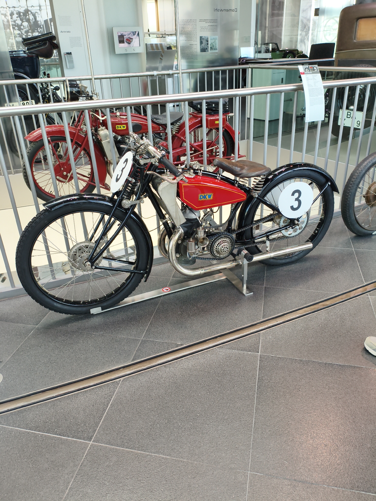 An antique red and black DKW motorcycle with the number 3 prominently displayed on a white circle on the rear fender is the central subject of this image. The motorcycle is positioned behind a metal barrier, likely in a museum or exhibition. The foreground shows the tiled floor of the exhibition space. To the left, another red motorcycle is partially visible. In the background, a display case with text and images, and part of a vintage car are visible. The overall impression is that of a historical exhibit within a public building. The context of Ingolstadt, Germany is provided, suggesting this museum or exhibition is located there.