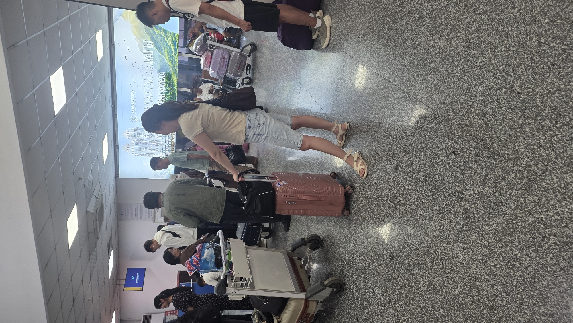 The image depicts a scene at an airport, likely in Nariman, Kyrgyzstan, judging by the signage. Several people are present, appearing to be waiting in line or moving through the terminal. In the foreground, a woman stands with a pink suitcase and a black duffel bag. To her left, a luggage cart holds several bags. In the background, a large digital billboard displays an image of buildings and text, which is partially obscured. The flooring is a speckled gray material, and the ceiling features fluorescent lighting panels. People are dressed in casual attire, and various pieces of luggage are visible throughout the scene, suggesting a travel context.