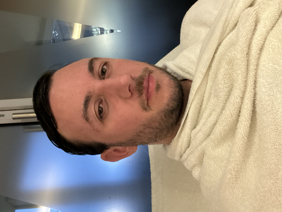 A man with short, dark hair and a beard is wrapped in a white towel. He faces the camera with a slight smile. Behind him, a gray wall is visible with a triangular-shaped reflective object mounted on it. The man's skin tone is fair, and he has light brown eyes. He appears relaxed and comfortable, possibly after a shower or spa treatment.
