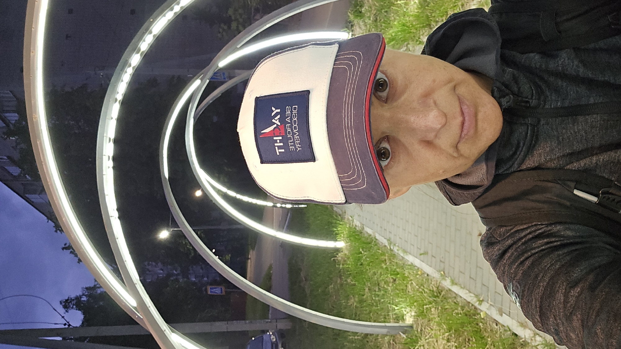 A first-person, medium shot captures a woman wearing a dark grey hooded sweatshirt and a white and blue baseball cap with the text "TH-AY SEA ROUTE DISCOVERY" visible on the front. The woman is standing on a brick pathway in what appears to be a park or pedestrian area illuminated by arched, white LED lights. The background shows a tree-lined street with a building and a street lamp. The time of day is dusk or evening. The woman is looking directly at the camera with a neutral expression. The overall scene is urban, with elements of greenery and artificial lighting.