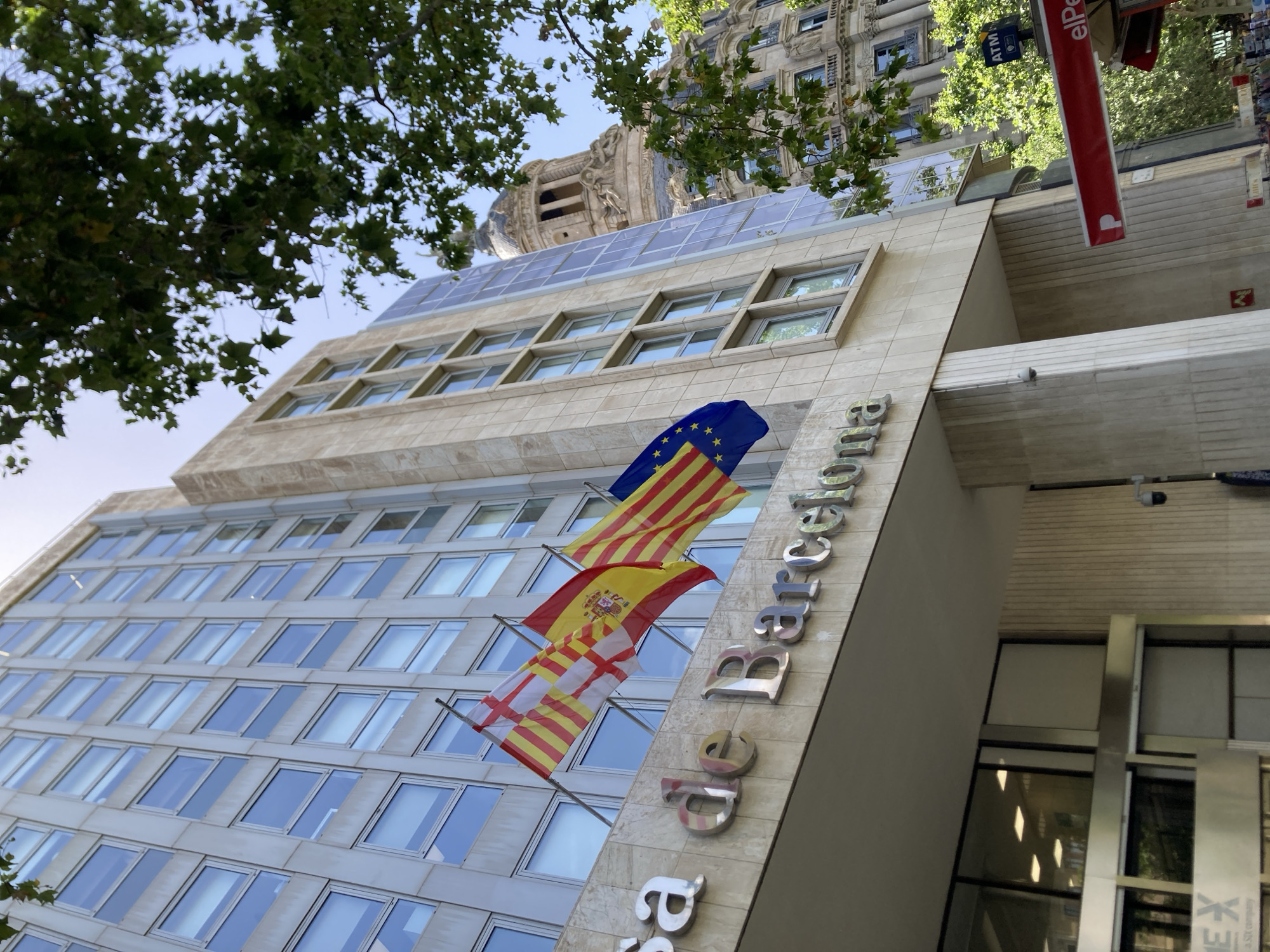 This media file depicts the exterior of a modern office building in Barcelona, Spain. The building's facade is constructed with light-colored stone and features numerous rectangular windows, suggesting multiple floors. The top of the building is equipped with solar panels, indicating a focus on sustainability.
Several flags are prominently displayed on the building, including the flag of Spain, the flag of the European Union, and the flag of Catalonia. These flags are attached to poles extending from the facade.
The building's name is visible in large, metallic letters on the side of the building, spelling out "de Barcelona". This inscription, along with the Catalan flag, strongly suggests the building is located in Barcelona.
In the foreground, large green tree branches with leaves partially obscure the view of the building, creating a contrast between nature and the urban environment.
In the background, an older, ornate building with intricate architectural details and a domed structure can be seen, suggesting a mix of architectural styles in the vicinity. A red sign with white lettering, which appears to be an advertisement or directional sign, is also visible. The time of day appears to be daytime, with bright lighting, and the weather seems fair. No people are visible in the image.