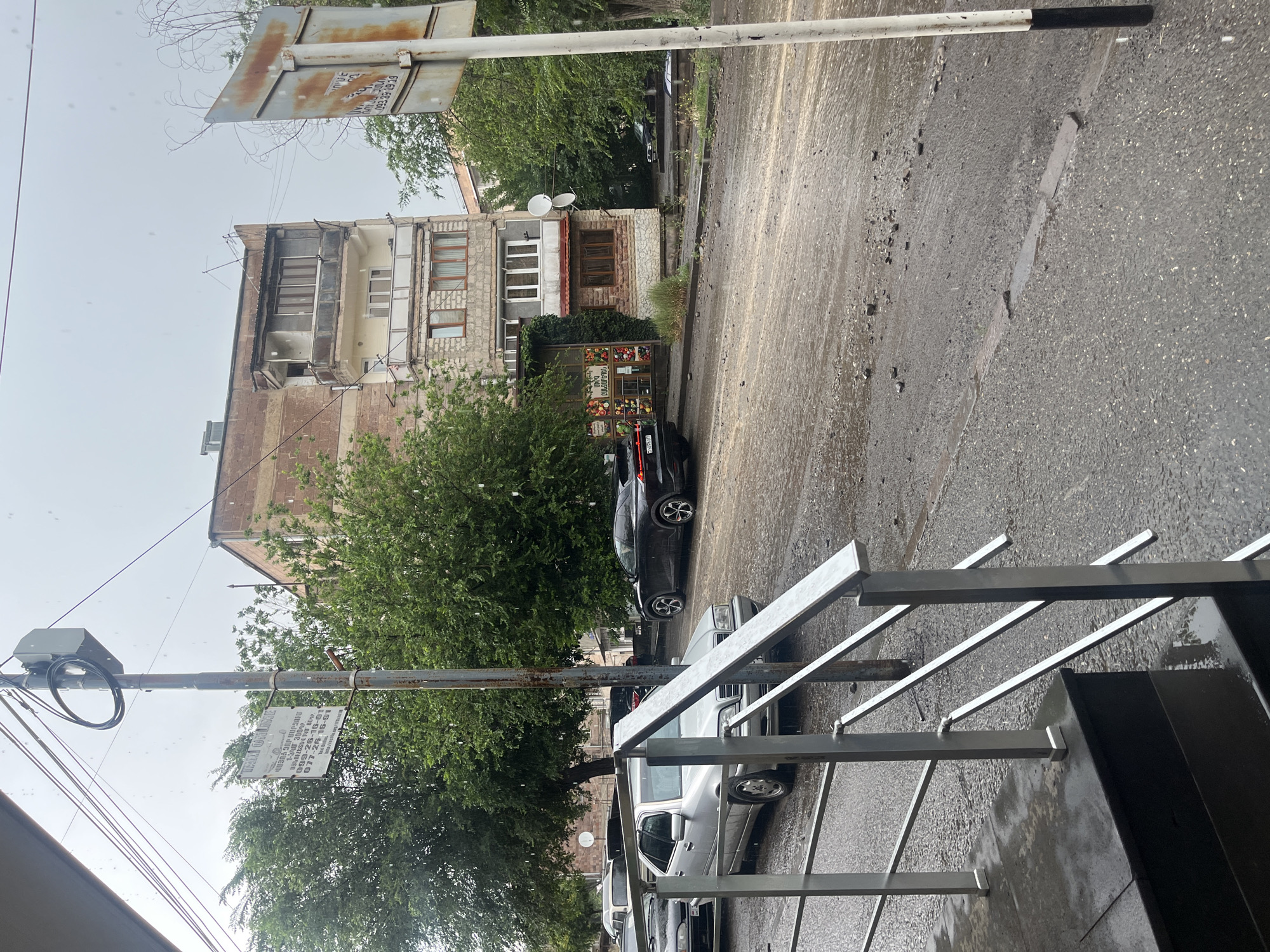 The image shows a street scene in an urban environment, likely during rain, as indicated by the falling droplets and wet surfaces. A multi-story building with balconies and brickwork stands in the background. Several cars are parked or driving on the wet asphalt road, including a silver sedan and a dark-colored SUV. A light pole with a sign attached is visible in the foreground, and another rusted sign is to the right. Trees with green foliage line the street. The foreground shows a set of metal railings and steps leading down. The road appears somewhat rough, with debris scattered on its surface. The sky is overcast, suggesting a gloomy or rainy day.