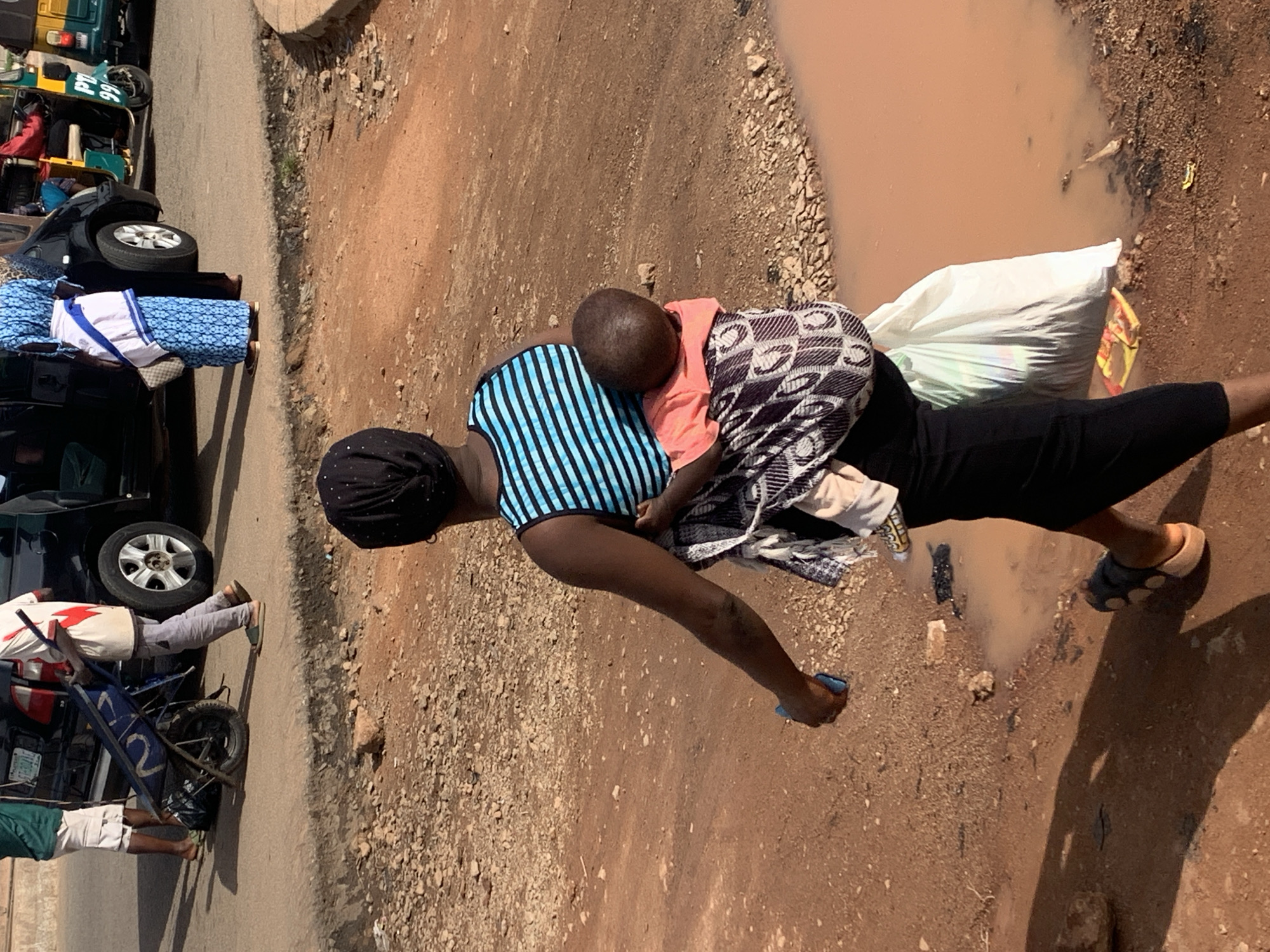 A woman in a black headscarf and a blue and black striped tank top carries a baby on her back, wrapped in a patterned cloth. She wears a long black skirt and is holding a blue object in her left hand. She is walking past a large puddle of muddy water on a dirt road. In the background, other people and vehicles, including a black car and a yellow auto-rickshaw, are visible on a paved road. The scene appears to be in Gura Top, Nigeria.
