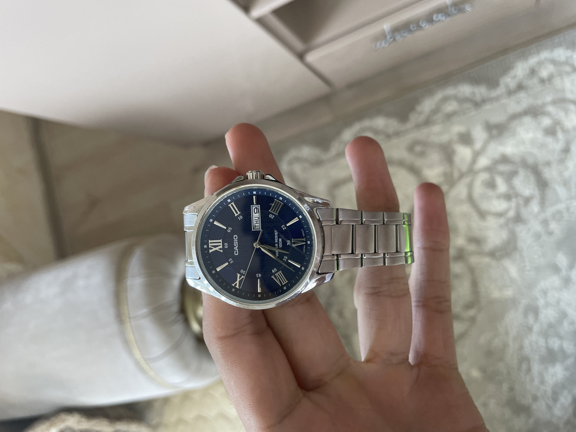 A close-up, eye-level shot captures a light-skinned hand holding a Casio wristwatch indoors in Ihtiman, Bulgaria. The watch features a silver-toned metal case and a matching link bracelet. Its dark blue dial is prominent, displaying a combination of Roman numerals (XII, IX, VIII) and index markers for the hours, along with smaller Arabic numerals (05, 10, 20, 25, 30, 35, 40, 45, 50, 55, 60) for the minute track. The day and date complications are visible, showing "THU 9". The watch's hands indicate a time of approximately 3:48.
Notable text on the watch face includes "CASIO" below the XII marker, and "WATER RESIST 100M" and "JAPAN MOVT" above the VI marker. The background is softly blurred but reveals light, neutral tones. To the left, a large, cylindrical or rounded beige object is partially visible. To the right and bottom, a patterned, light-colored surface, possibly a rug or upholstery, can be seen. The top portion of the background shows indistinct light-colored walls or ceiling, with some faint, reflective marks suggesting a glossy surface or distant object. The lighting is bright and even, consistent with an indoor daytime setting. The hand holding the watch is positioned centrally, presenting the watch clearly to the viewer.