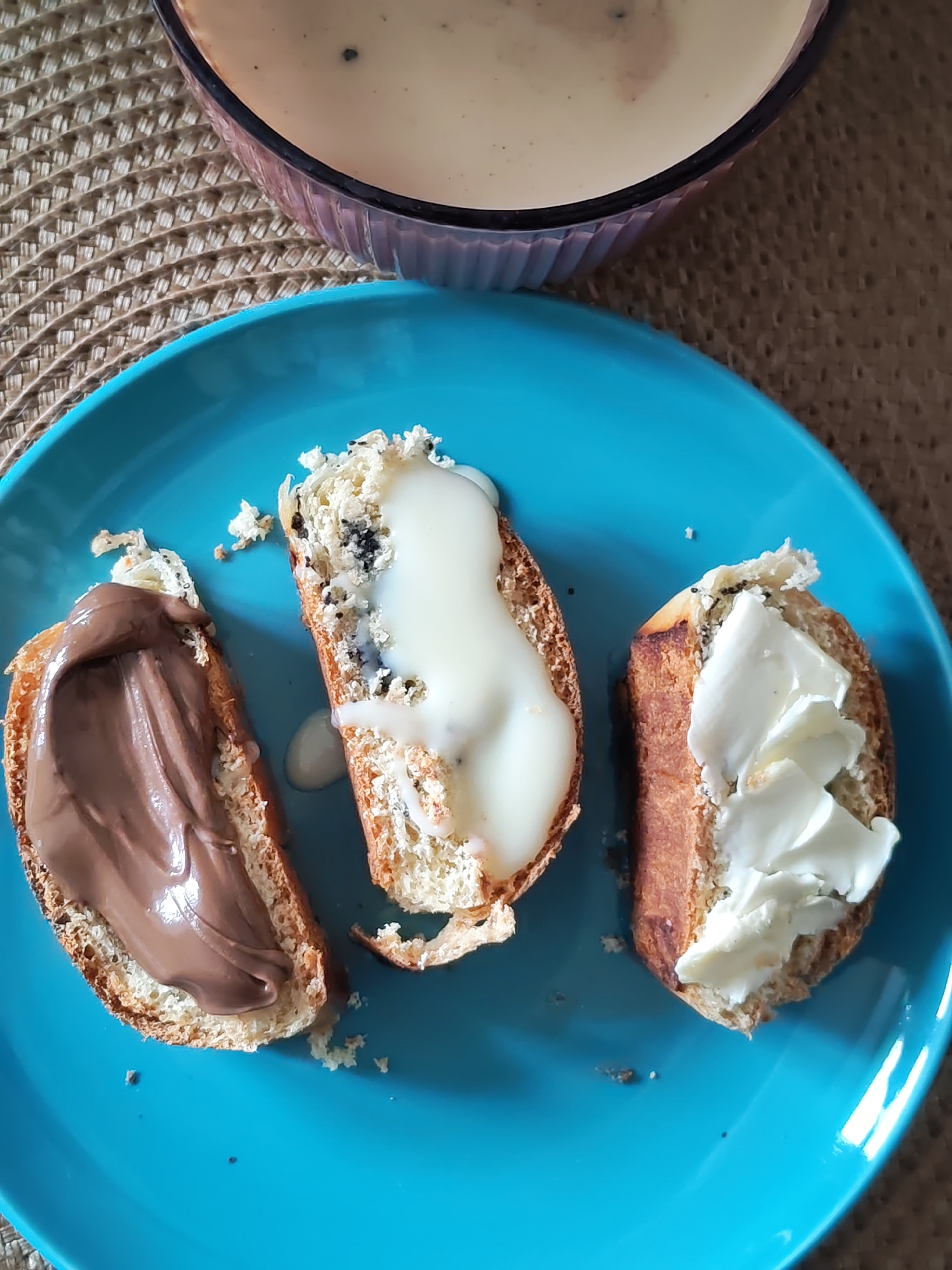 The image shows a light blue plate with three slices of bread, each topped with a different spread. One slice has a chocolate-colored spread, another is covered with a white spread that has some black specks underneath, and the third slice is topped with a white, creamy spread. There are crumbs scattered around the bread on the plate. A bowl of light-colored liquid is partially visible in the upper part of the image, sitting on a woven placemat. There is no information suggesting this image was taken in Izhevsk, Russia.