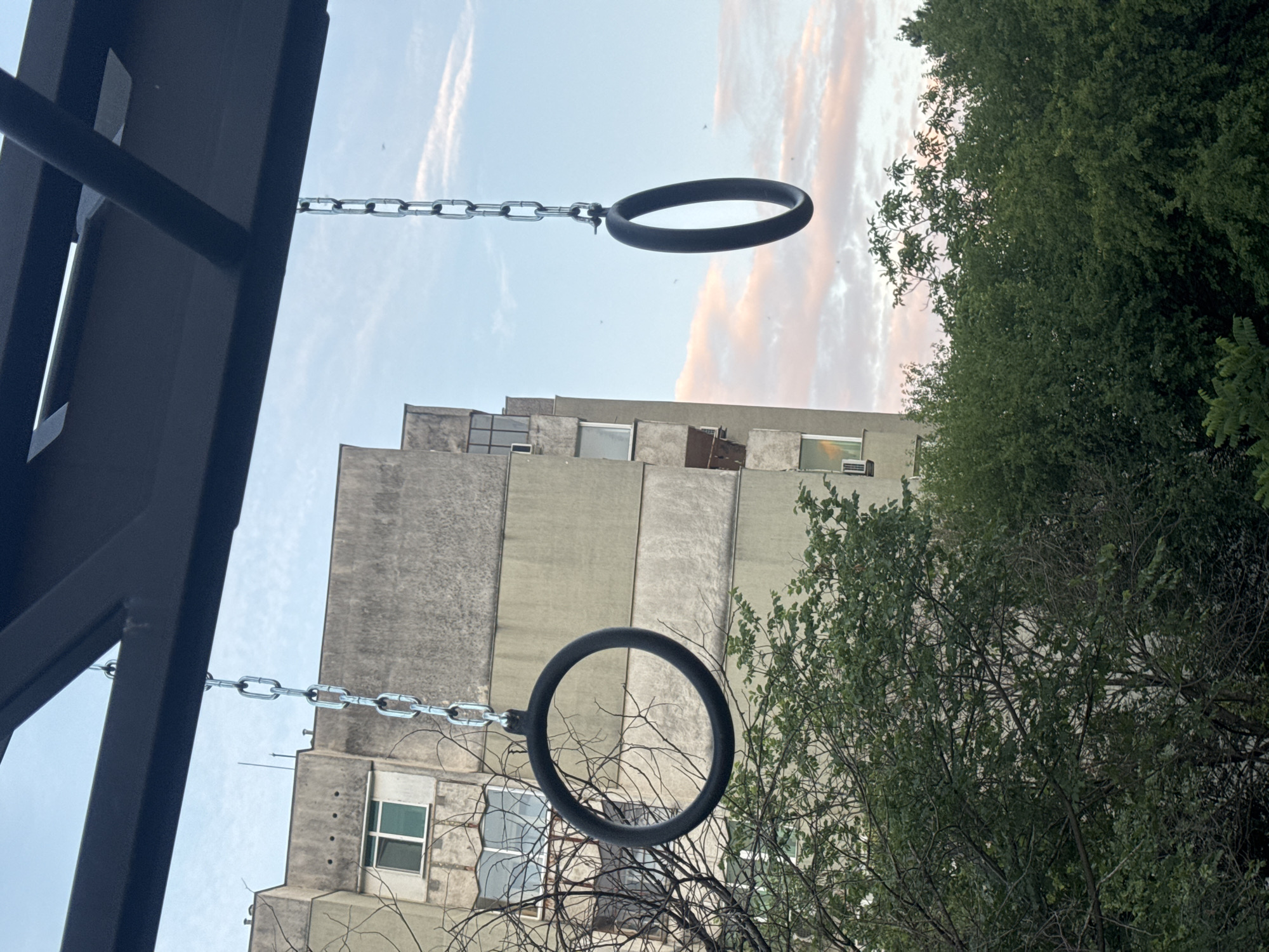 The image depicts a close-up view of two gymnastic rings hanging from chains, suspended beneath a dark metal structure. In the background, a multi-story building with light-colored facade and numerous windows is visible, partially obscured by lush green trees. The sky above is a pale blue with streaks of pink and white clouds, suggesting either sunrise or sunset. The scene appears to be an outdoor recreational area, likely a playground or a park within the city of Stara Zagora, Bulgaria. There are no people or animals present in the image, and no specific actions or interactions are taking place. The focus is on the gymnastic rings and their immediate surroundings.