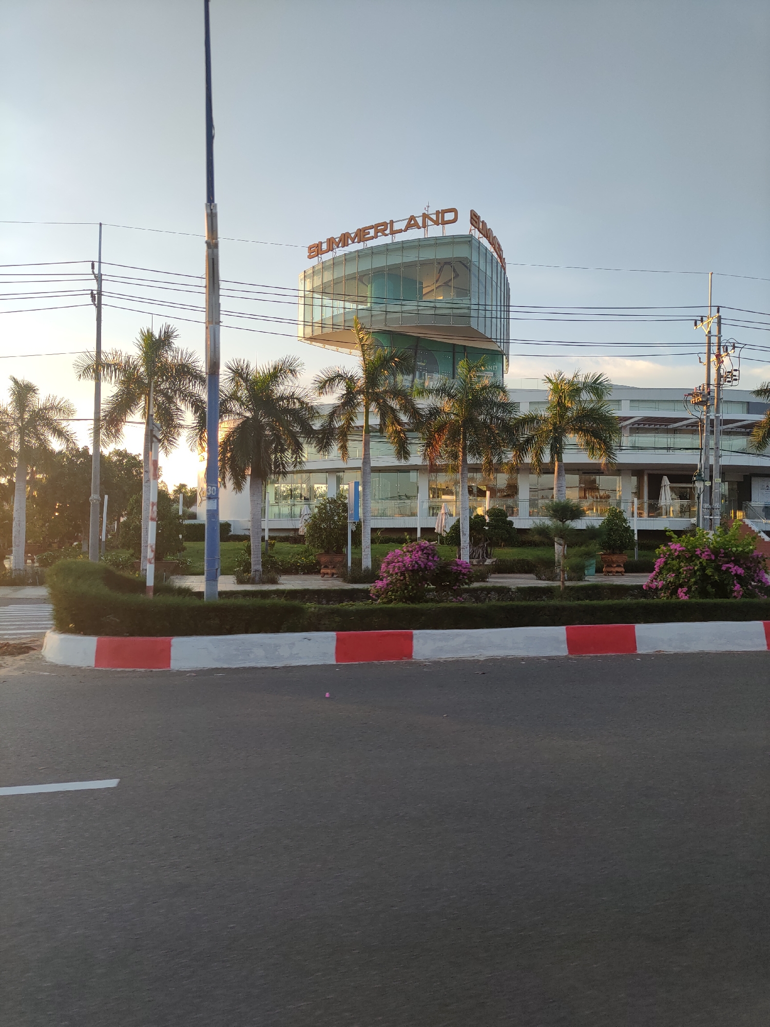 The image captures a street view of the Summerland building in Phu Thuy Ward, Vietnam. The building features a modern architectural design with a glass facade and orange signage that reads "SUMMERLAND". In front of the building, there are several palm trees and lush green landscaping, including flowering bushes in vibrant pink. A street with a crosswalk is visible in the foreground, bordered by a red and white curb. Utility poles and power lines run across the top of the frame. The sky is clear with soft, diffused sunlight, suggesting it is either early morning or late afternoon. No people are visible in the frame.