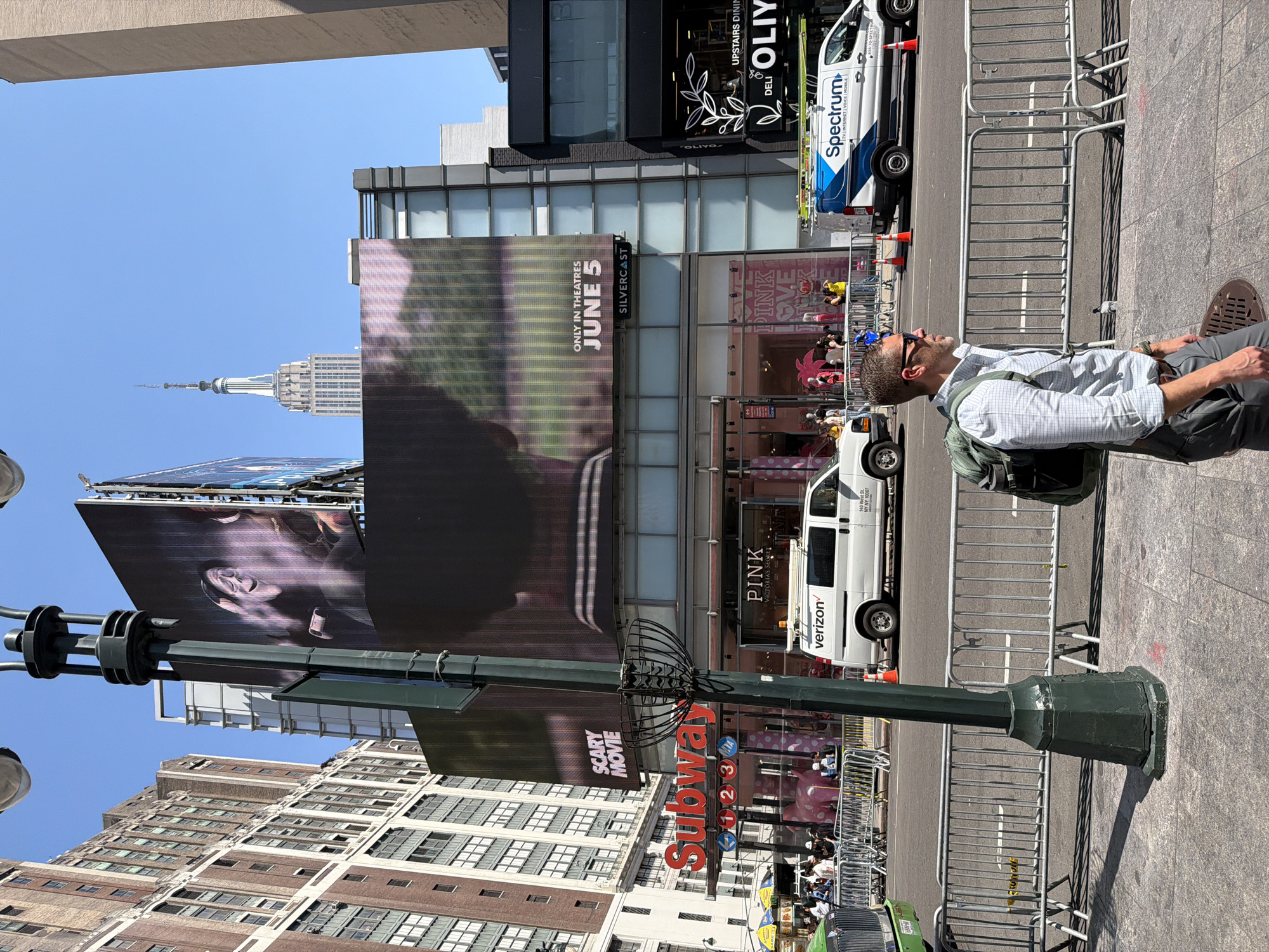 The image shows a man sitting on a sidewalk in New York City, USA, in what appears to be Times Square. He is wearing sunglasses, a button-down shirt, gray pants, and a backpack. He is looking up at the billboards and signs in the area. Above him, one billboard advertises a movie in theaters June 5th, and another advertises "Scary Movie." A third billboard shows a woman's face. Visible brands are "Subway" and "Verizon," "Spectrum," and a "PINK" Victoria's Secret store. The Empire State Building can be seen in the background. A Spectrum van, a Verizon van, and a green Taxi cab are parked on the street. Metal barricades line the sidewalk. The sky is blue and clear.