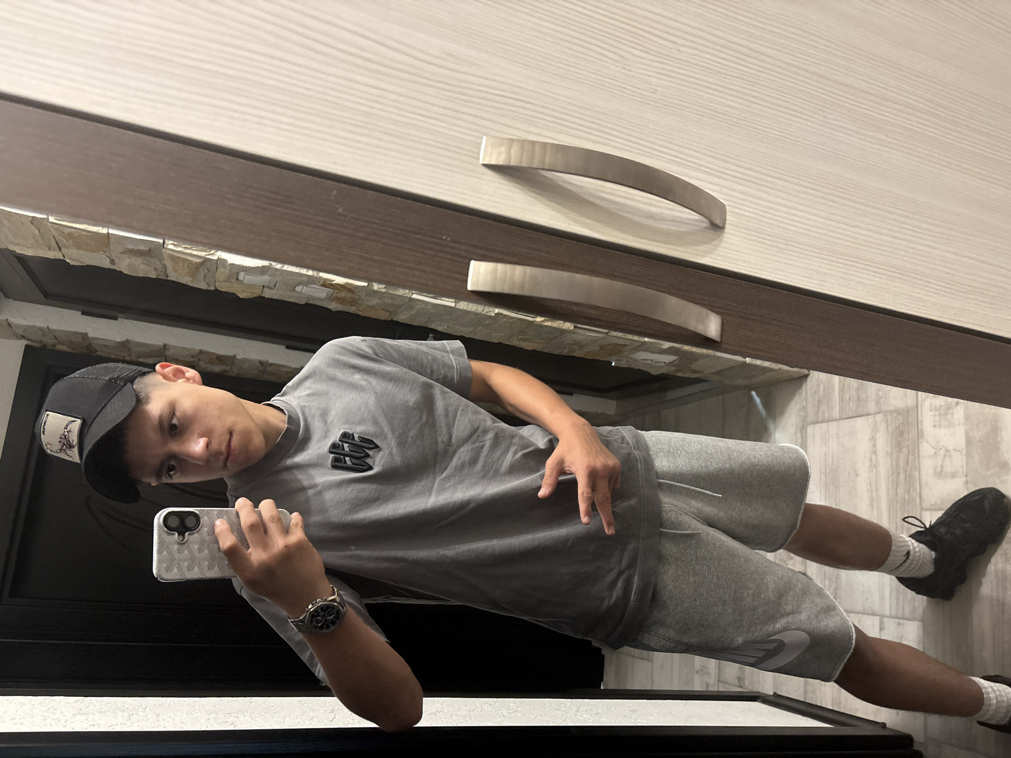A young man takes a selfie in a room with a cabinet or closet with horizontal grain and curved handles, trimmed with a stone-like texture. The floor has a light tile pattern. He is wearing a grey t-shirt with a logo, grey Nike shorts, black shoes with white socks, a black baseball cap, and a watch. The image is taken indoors and the exact location in Stara Zagora, Bulgaria, cannot be determined without additional contextual information.