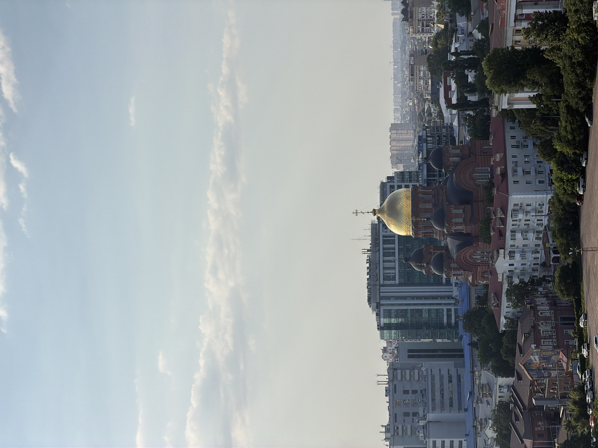 A high-angle medium shot captures a panoramic view of an urban landscape under a bright, cloudy sky. In the foreground, a large, red brick cathedral with multiple domes, the central one being gilded, dominates the scene. Surrounding the cathedral are various buildings, including residential apartments and modern office buildings with glass facades. A dense green canopy of trees partially obscures some of the lower structures. In the middle ground and background, the city extends with numerous buildings and a hazy skyline. Several cars are visible parked in a lot in the lower left and lower right of the frame. The overall lighting suggests it is daytime, possibly late afternoon due to the soft light.