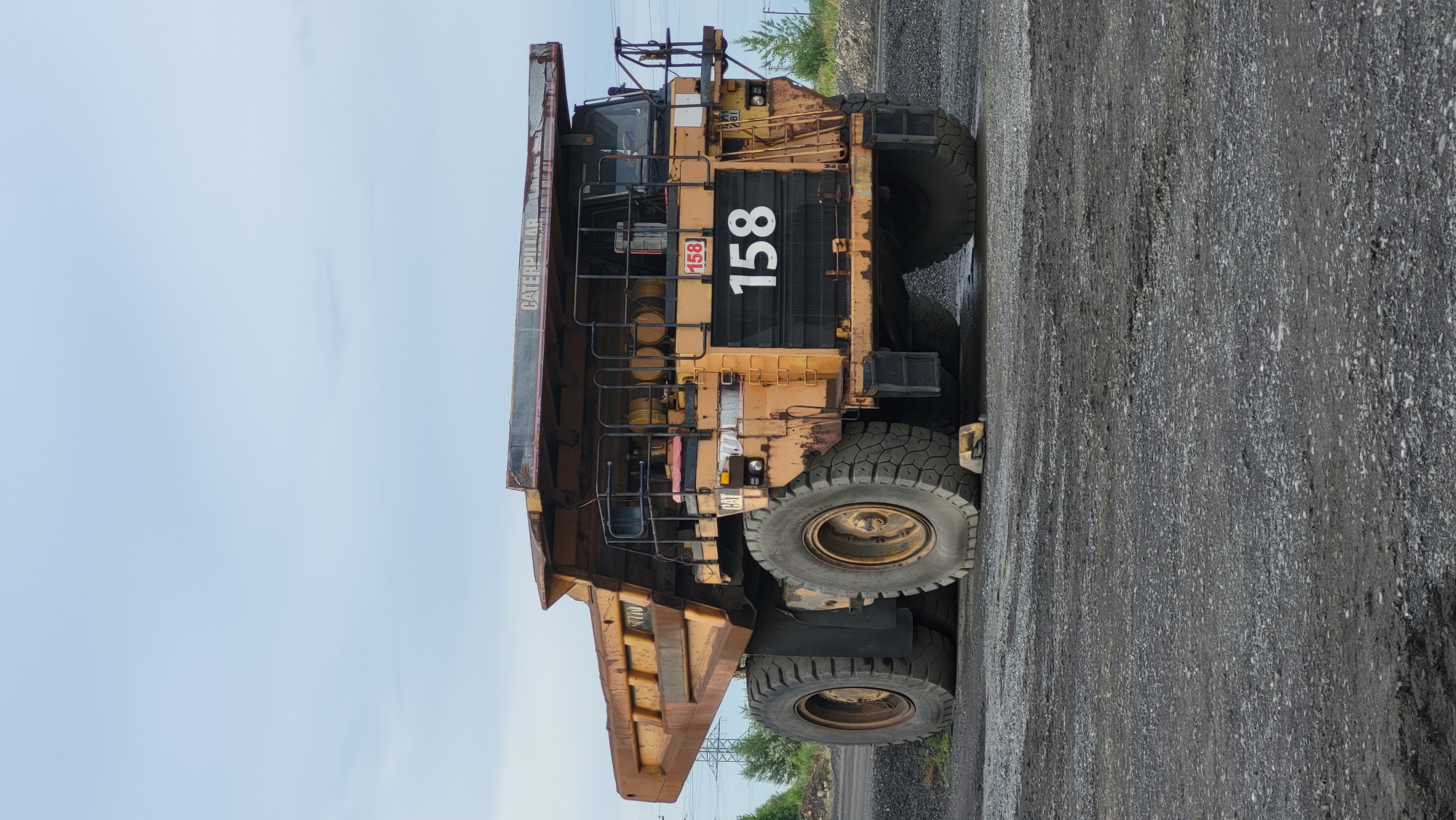 A large, yellow, rusty Caterpillar haul truck, numbered 158, is parked on a gravelly surface. The truck is massive, with enormous tires. The bed of the truck is raised and appears to be empty. The cab of the truck is visible at the top right, with a dark tinted windshield. Above the truck, the sky is overcast and pale blue. The background consists of a few scattered trees and power lines. The scene appears to be in an industrial area or a mine. The location of Voloshyne, Ukraine is not discernible from the image itself.