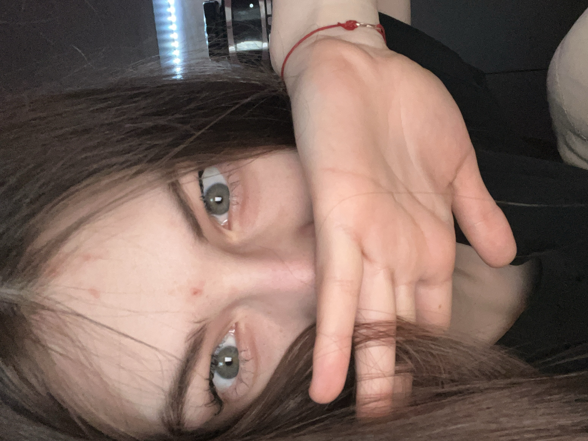 A close-up, angled shot of a person lying down, with their face and hand in focus. The person has light skin, brown hair, and grey eyes. Their hand is positioned near their face, with some hair tangled through the fingers. A red bracelet adorns their wrist.
The background is dark and blurred, with hints of a black object and a blurry reflection.
There are no discernible elements to establish a location in Saint Petersburg, Russia.