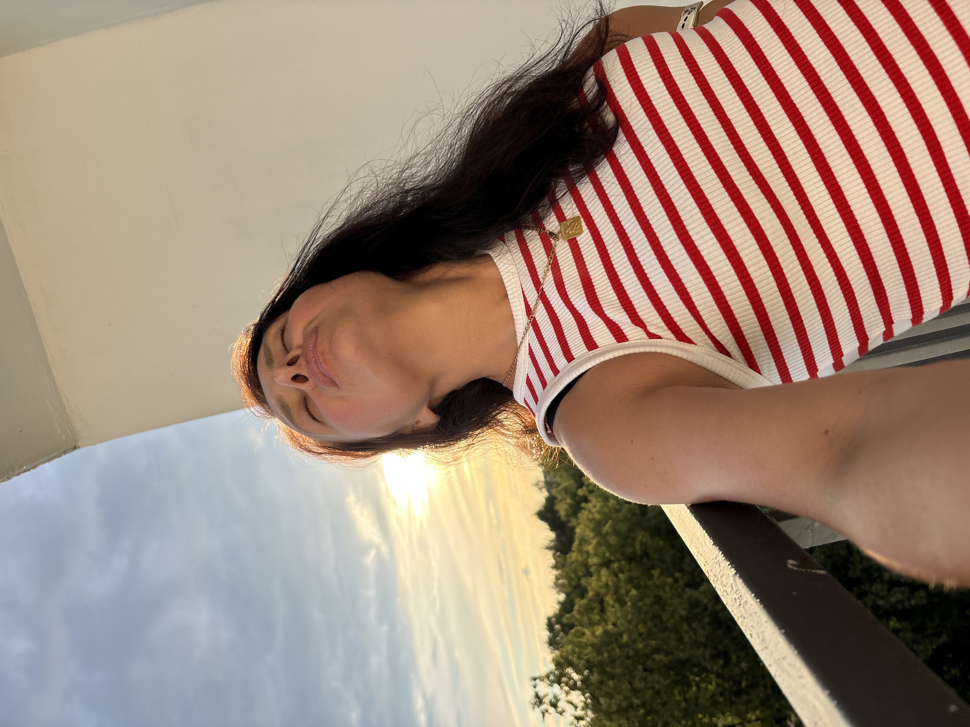 An outdoor selfie taken from a low angle shows a woman with her eyes closed and head tilted back, facing a sunset. She has long, dark hair and is wearing a red and white striped tank top and a gold pendant necklace. Behind her, the sky is a mix of blue and grey clouds, with the sun visible as a bright light. Lush green trees are visible in the background, along with a balcony railing in the foreground. The image does not contain information about the location in Chaikovsky, Russia.