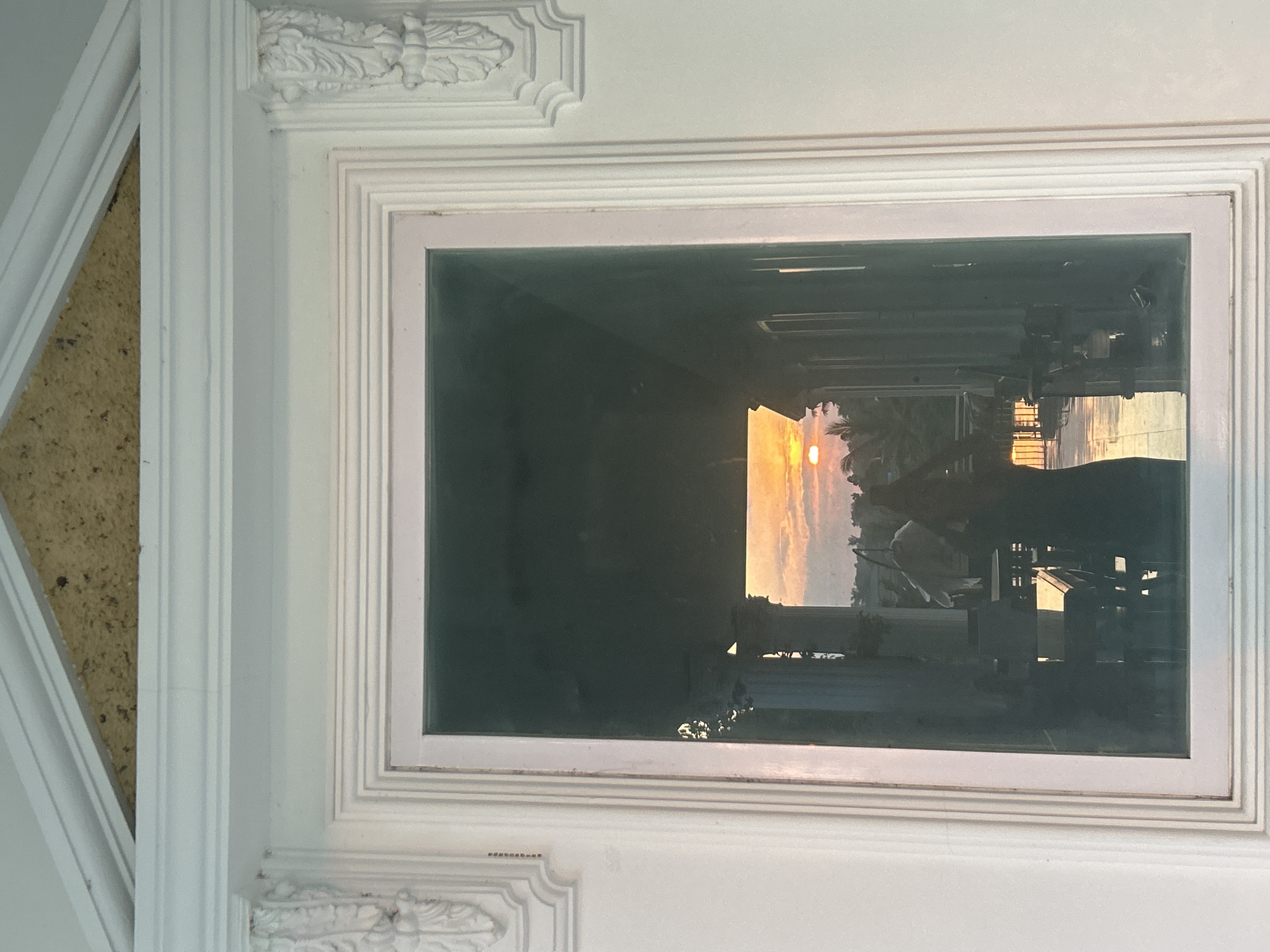 The image presents a head-on view of a white, ornate architectural frame, likely a large window or doorway, set within a light-colored wall. Above the frame, a roughly triangular gap reveals an unfinished ceiling or wall section composed of a coarse, yellowish-brown, granular material. The frame itself features multi-layered molding and decorative corbels on both vertical sides, carved with acanthus leaf motifs. The large glass pane within the frame reflects a complex scene.
Through the reflected glass, a sunset is visible with the sun as a distinct, bright orange disc positioned low on the horizon. The sky is filled with orange and pink hues, indicative of dusk. In the foreground of the reflection, two human figures are present: one on the left, partially obscured, appears to be wearing or holding something light-colored; the other on the right is a darker silhouette, possibly wearing a dress, standing further back. Between these figures and the sunset, architectural columns are reflected, suggesting an indoor or covered outdoor space beyond the immediate view. Furniture, including what appear to be tables and chairs, is also reflected in the mid-ground. Beyond the columns and furniture, silhouetted tropical trees, possibly palm trees, are visible against the sunset sky. The reflection of the sunset illuminates the floor or ground surface within the reflected scene, creating a warm glow. The overall scene depicts a view through a window, capturing both the exterior environment and reflections of an interior space at sunset in the city of Unknown, Unknown.