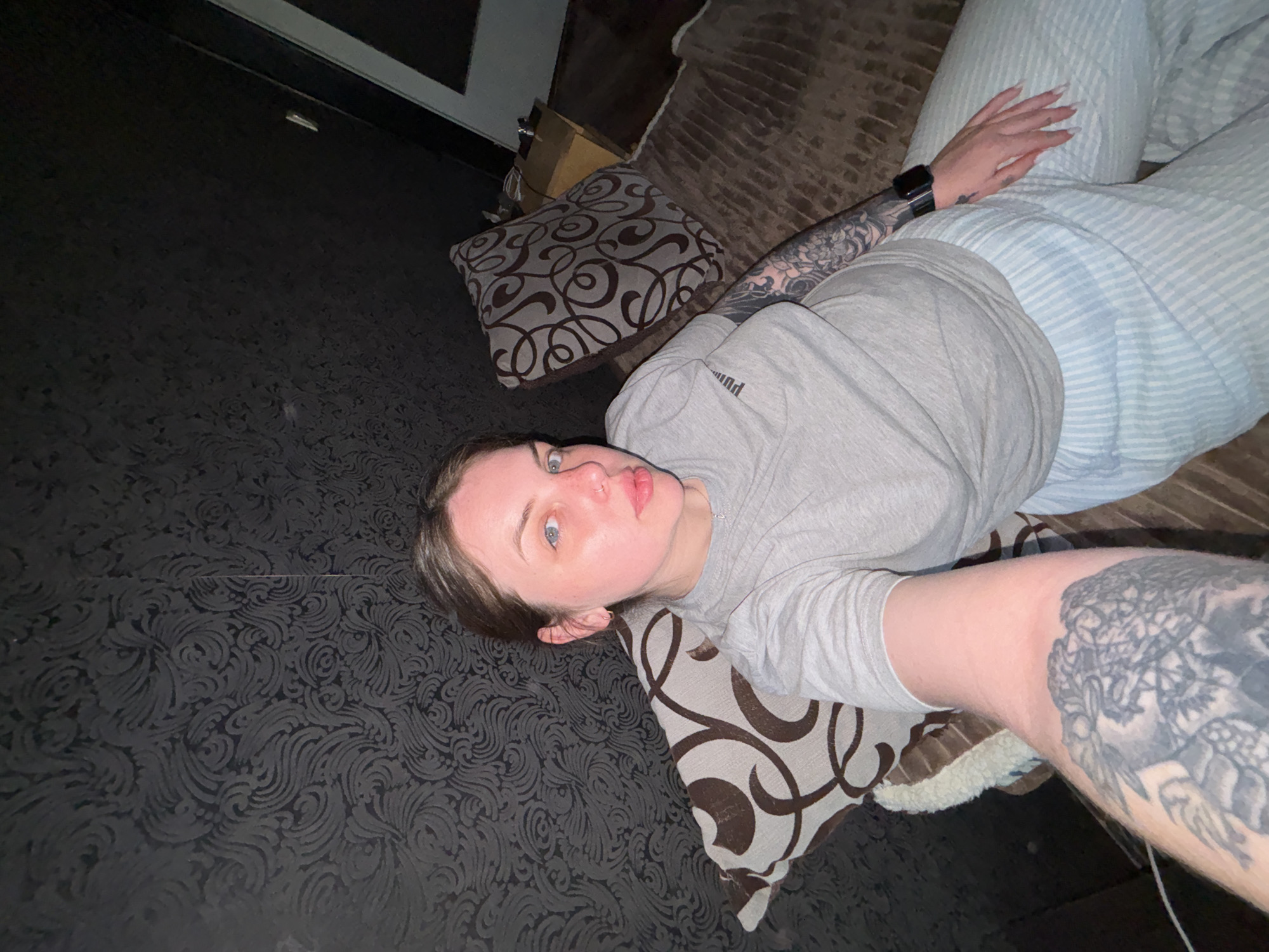 A young person is seen in an indoor setting, possibly a living room or bedroom, captured from a high-angle, tilted perspective. They are lying on their back, looking directly at the camera with wide blue eyes and a slightly pouting expression.
The person is wearing a light grey t-shirt, on which the brand name "PUMA" is visible on the upper left sleeve. They also wear light blue and white striped pajama-style pants. A dark smartwatch is visible on their left wrist, and they have a nose piercing. Both arms are extensively covered in dark tattoos, with intricate designs visible on the sleeves. Their right hand, with long, light-colored nails, rests on their midsection.
They are lying on a large, soft surface, likely a sofa or bed, which appears brown or dark brown. Next to them is a patterned throw pillow with a swirling brown and grey design. Behind this pillow, a cardboard box is partially visible. The background features a dark wall or floor covering with a subtle, embossed or flocked pattern of swirling motifs.
The lighting is somewhat dim, suggesting an indoor environment without bright natural light. The overall mood is casual and relaxed. While the image itself does not provide specific geographical cues, the context provided states the location is Chornomorskoe, Ukraine.